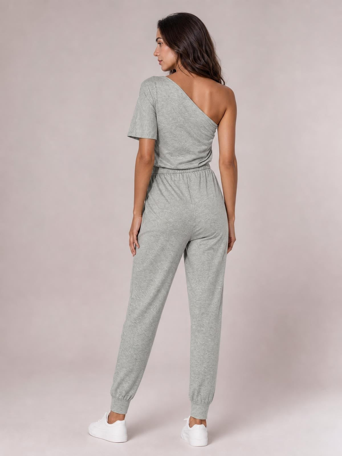 A model wears a gray single-shoulder short-sleeve jumpsuit with a relaxed jogger silhouette and elastic cuffs, shown from the back.