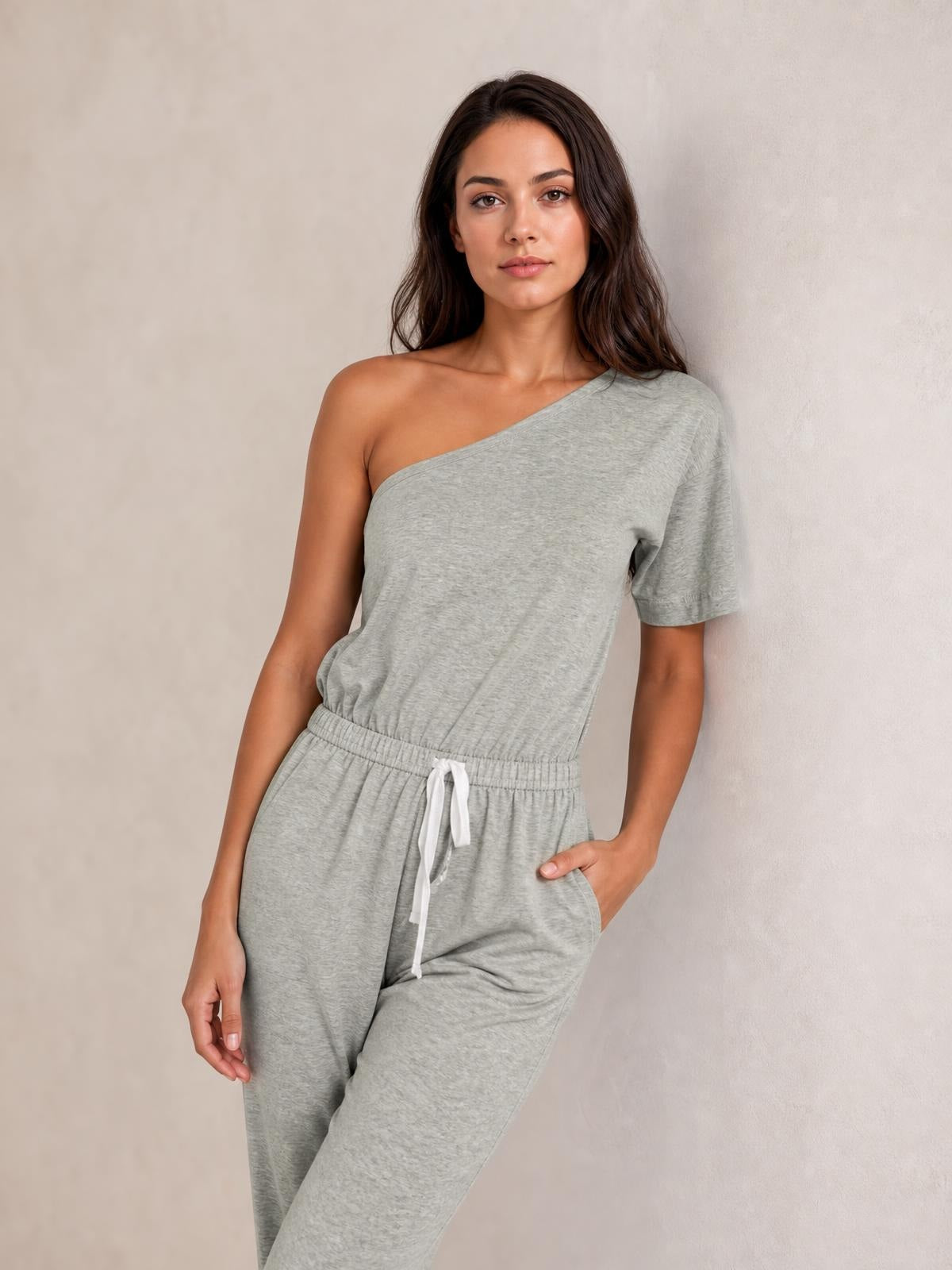 Model poses in a light gray single-shoulder short-sleeve jumpsuit with an elastic, tied drawstring waist and relaxed wide-leg silhouette.