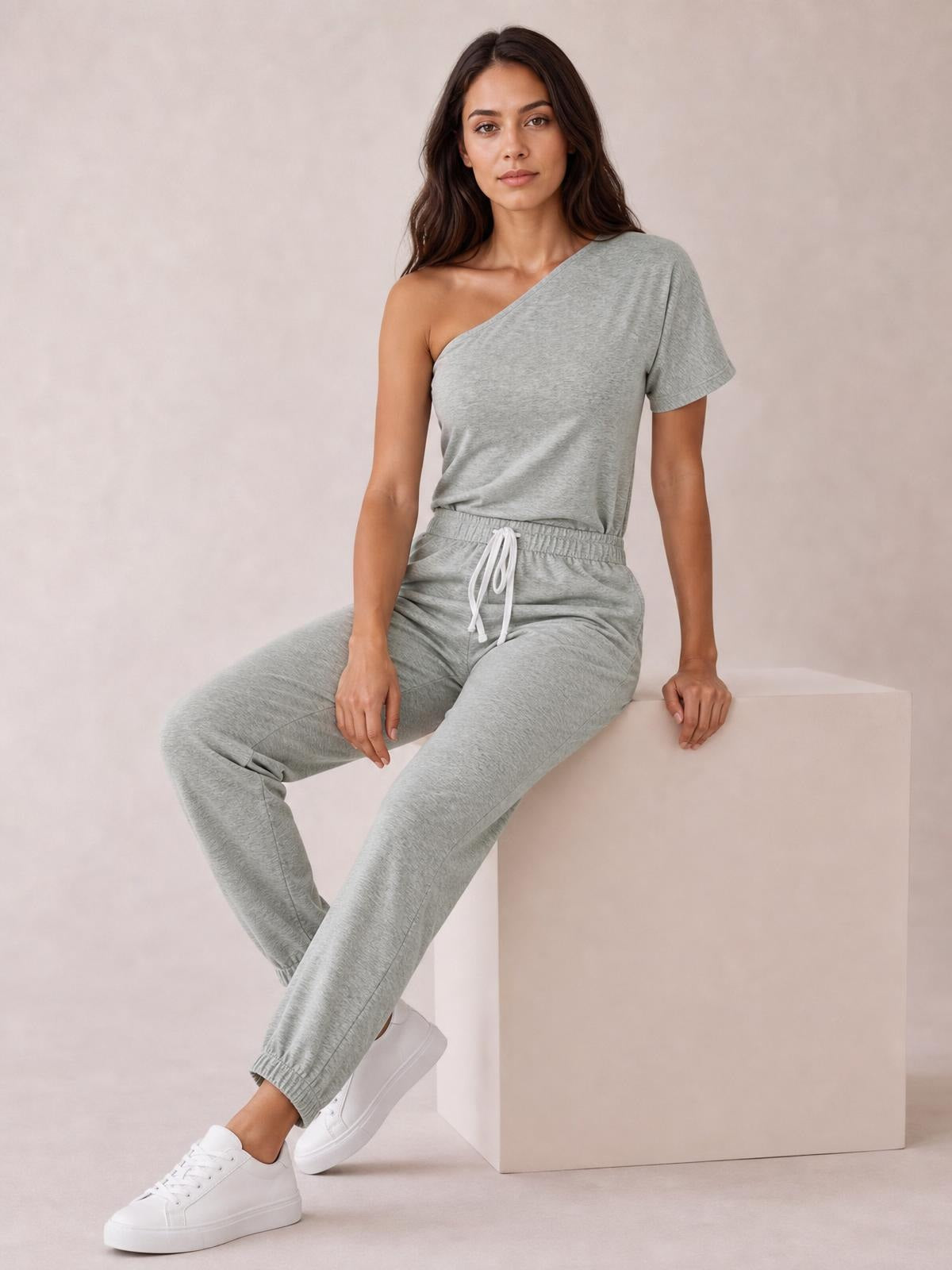 A model sits in a gray single-shoulder short-sleeve jumpsuit with a tied elastic waist, relaxed tapered legs, and a comfortable casual silhouette.