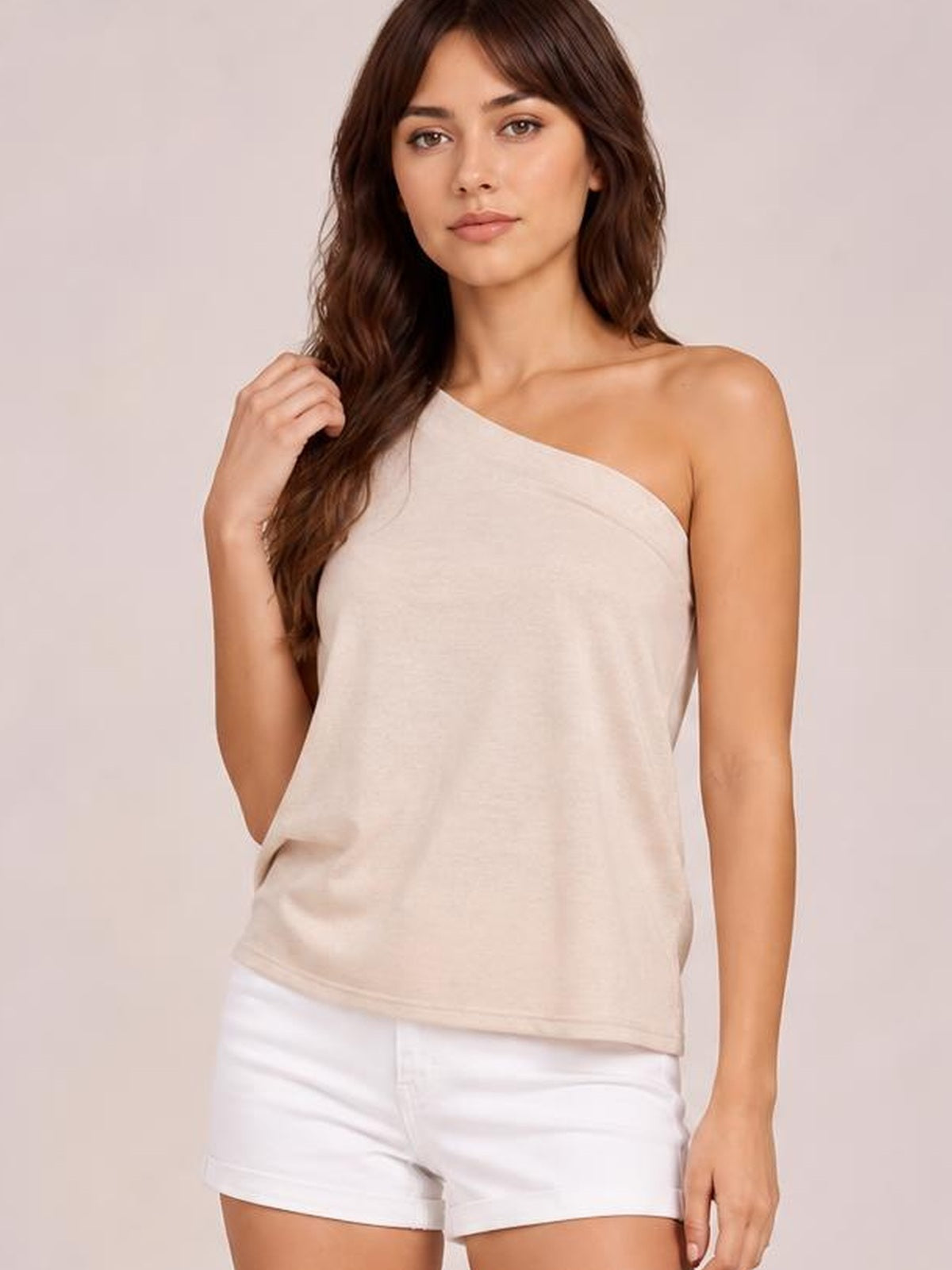 A model poses in a light beige single shoulder short sleeve top with an asymmetrical neckline, relaxed silhouette, and smooth drape while touching her hair.