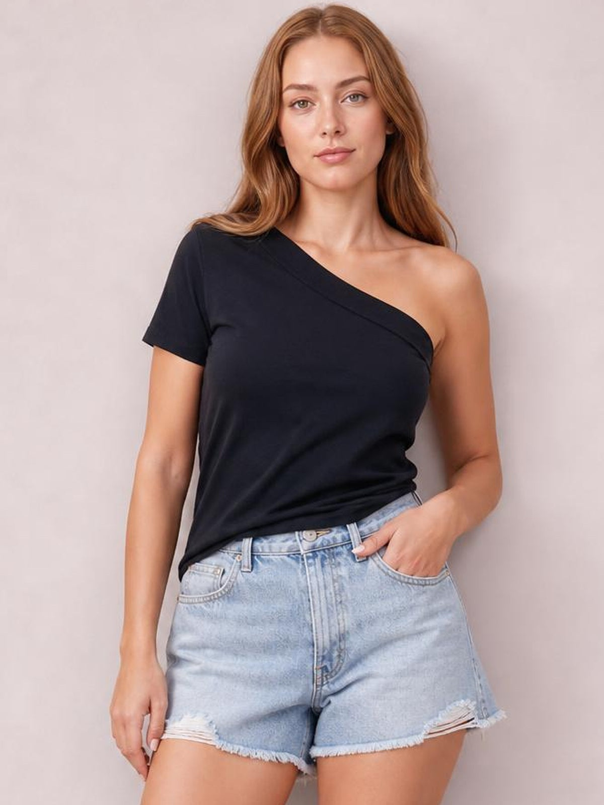 Model wearing a black asymmetrical single-shoulder short-sleeve top with a relaxed silhouette, paired with light-wash denim shorts and one hand in a pocket.