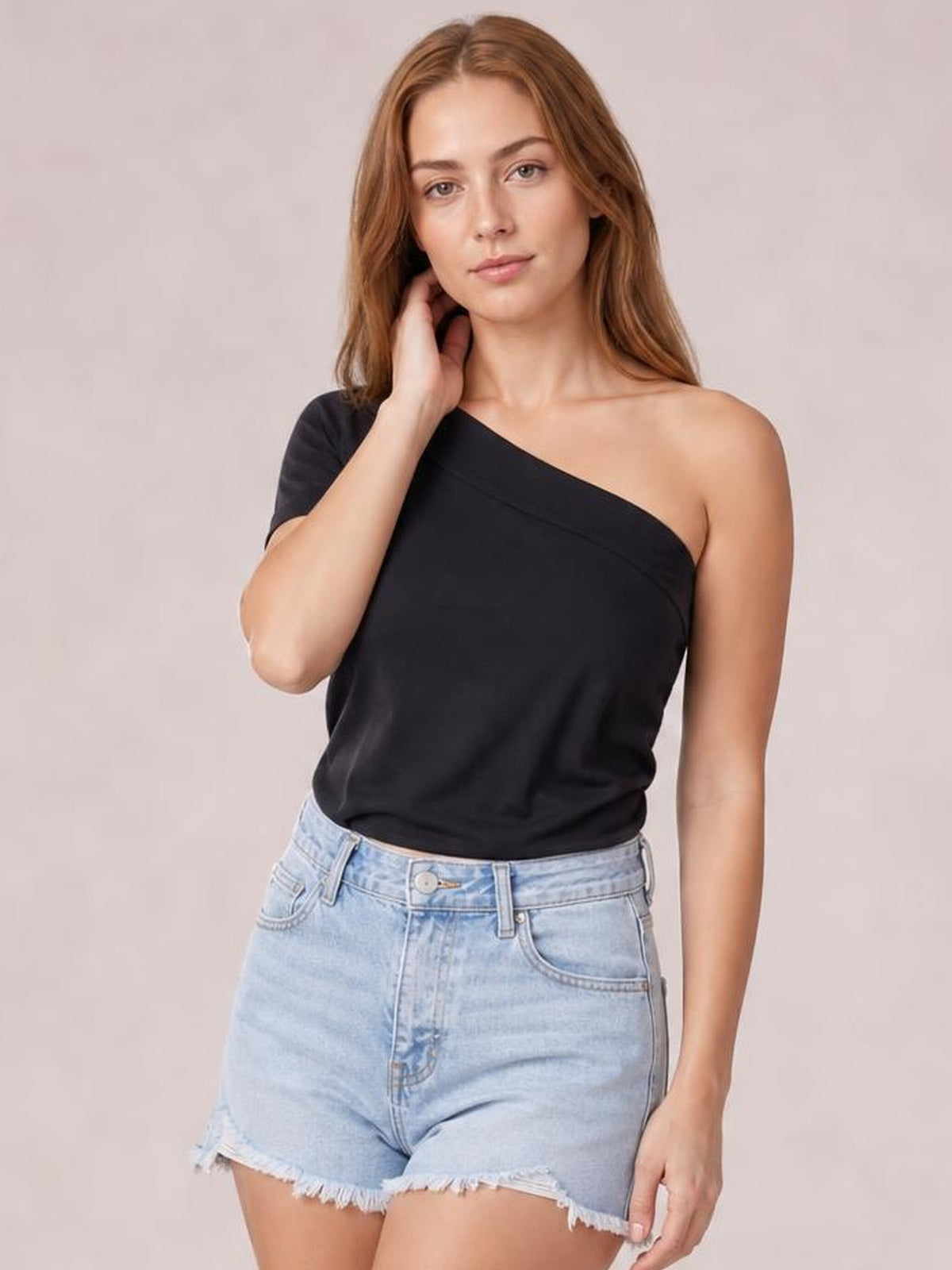 Model poses in a black single shoulder short sleeve top with an asymmetrical neckline and relaxed silhouette, paired with light-wash denim shorts.