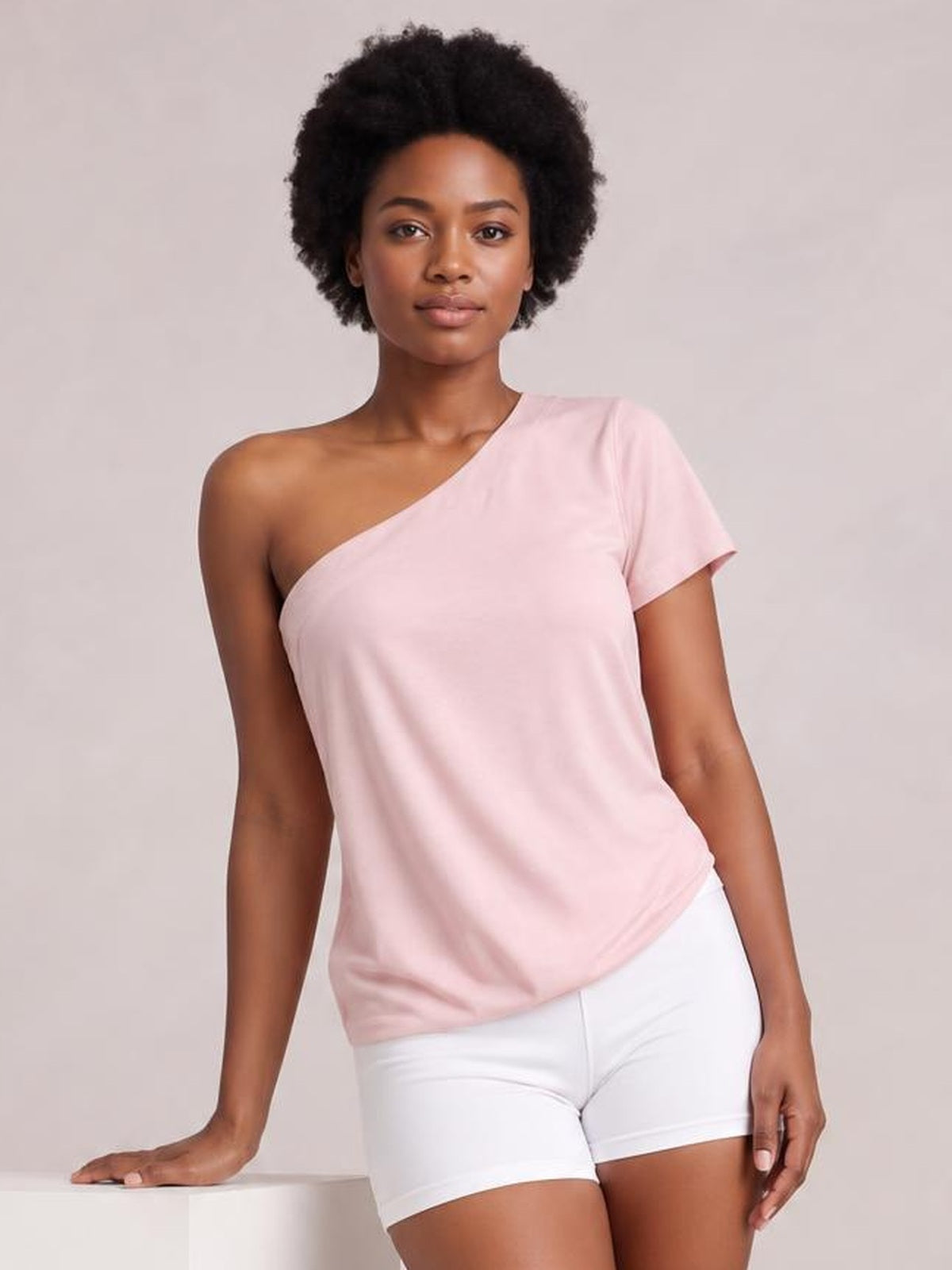 Model in a relaxed pose wearing a pale pink single shoulder short sleeve top with an asymmetrical neckline and relaxed straight silhouette.