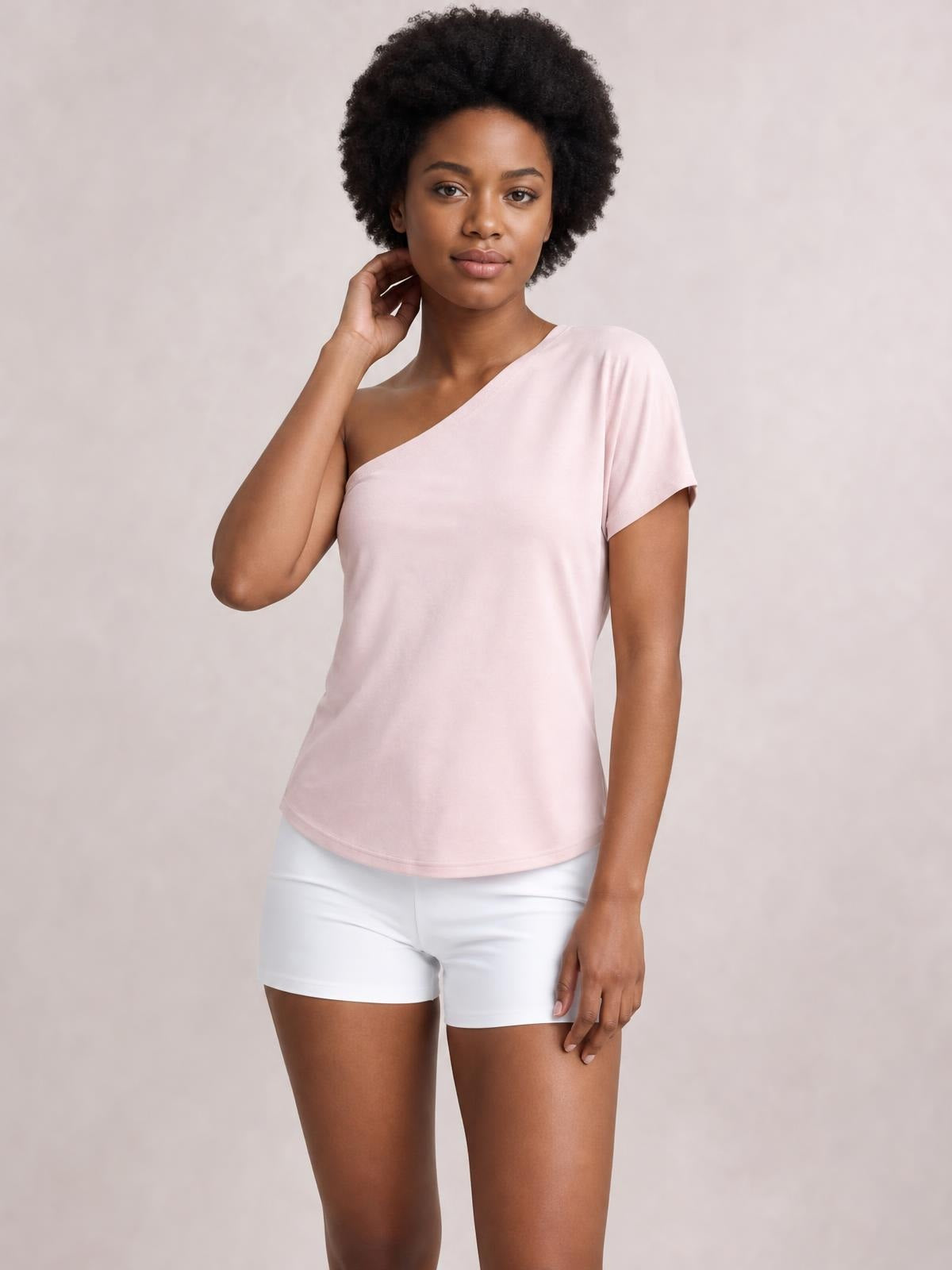 Model poses in a pale pink single shoulder short sleeve top with an asymmetrical neckline, relaxed silhouette, and curved hem.