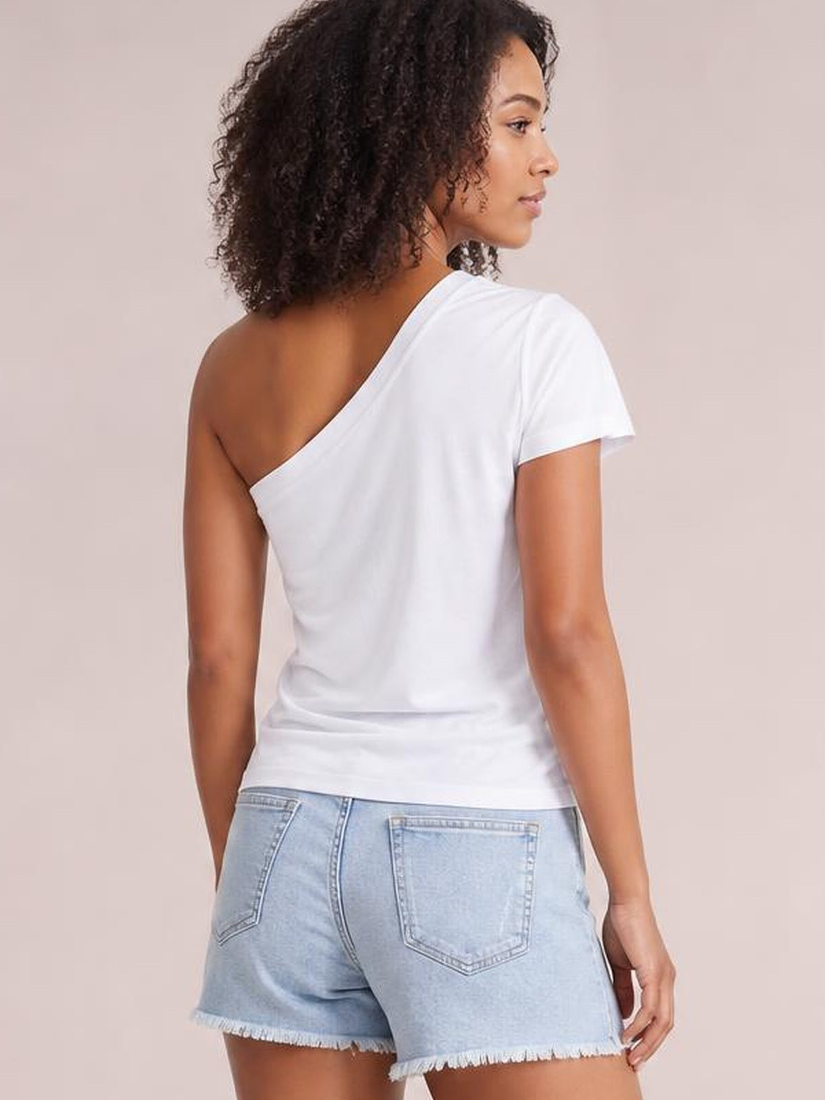 Model shown from behind wears a white asymmetrical single-shoulder short-sleeve top with a fitted silhouette and one exposed shoulder, standing with one hand at her side.