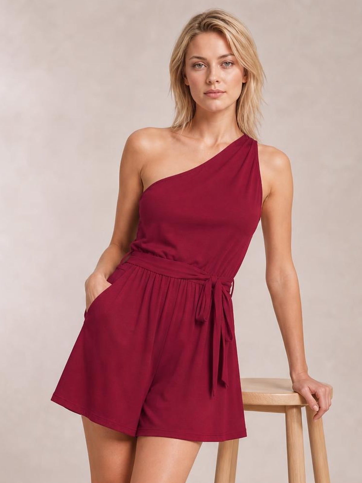 A model poses in a burgundy single-shoulder romper with a tie waist, relaxed shorts silhouette, and side pockets, resting one hand on a stool.