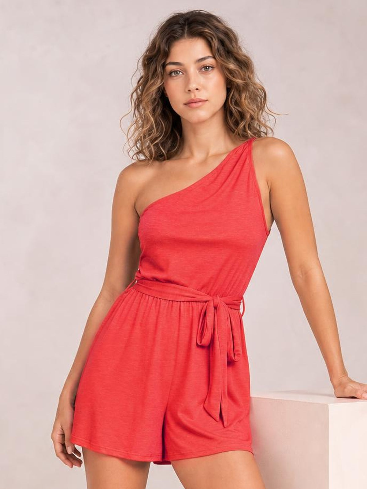 A model wears a coral single-shoulder romper with a relaxed silhouette, adjustable tie waist, and flowing shorts while leaning against a ledge.