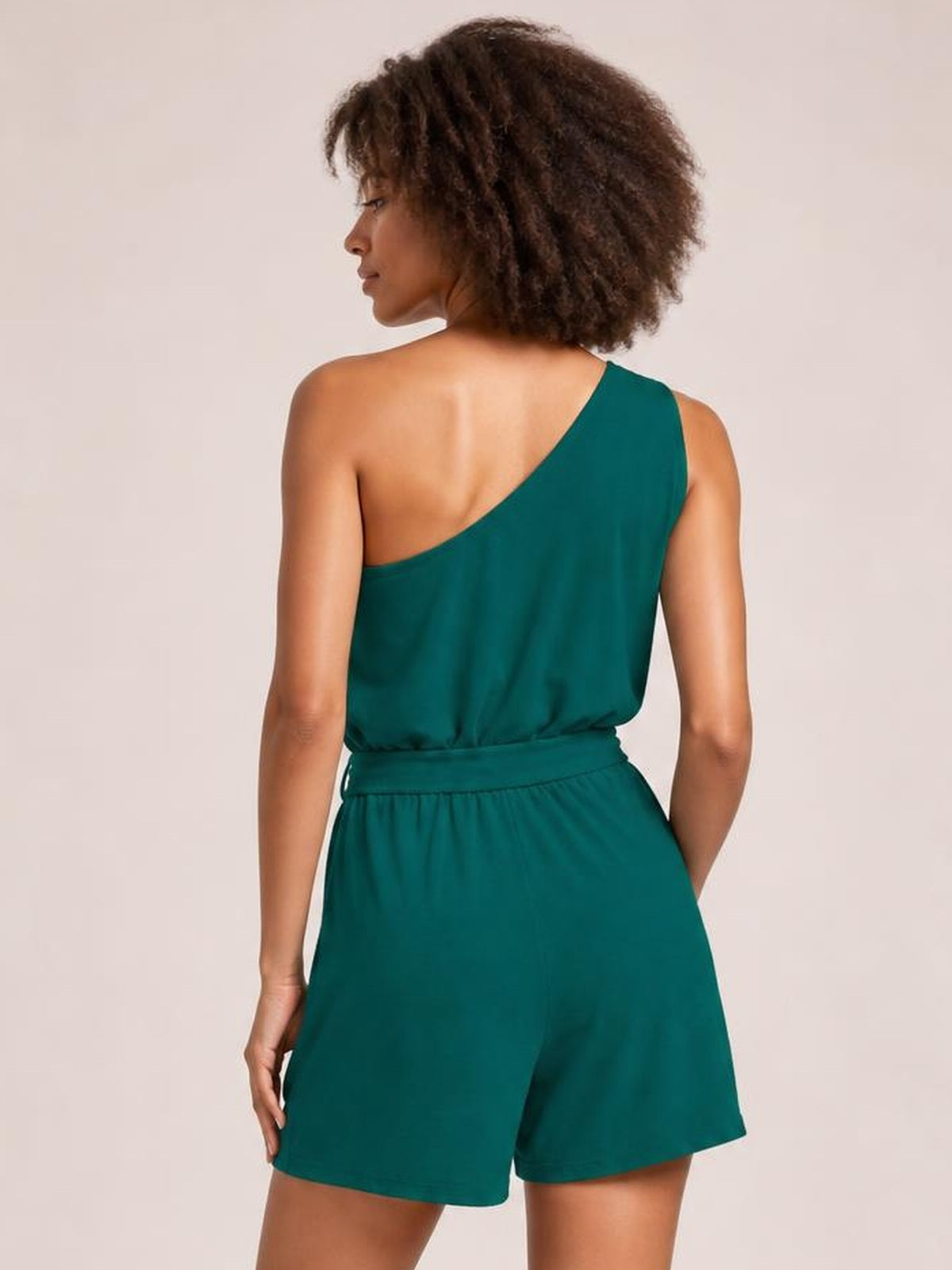 A model poses from the back in a teal single-shoulder tie waist romper with an asymmetric neckline, relaxed shorts, and a side pocket.