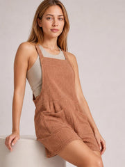 Model leans against a ledge wearing rust-brown sleeveless double gauze overalls with wide straps, a bib front, relaxed shorts, and large front pockets.