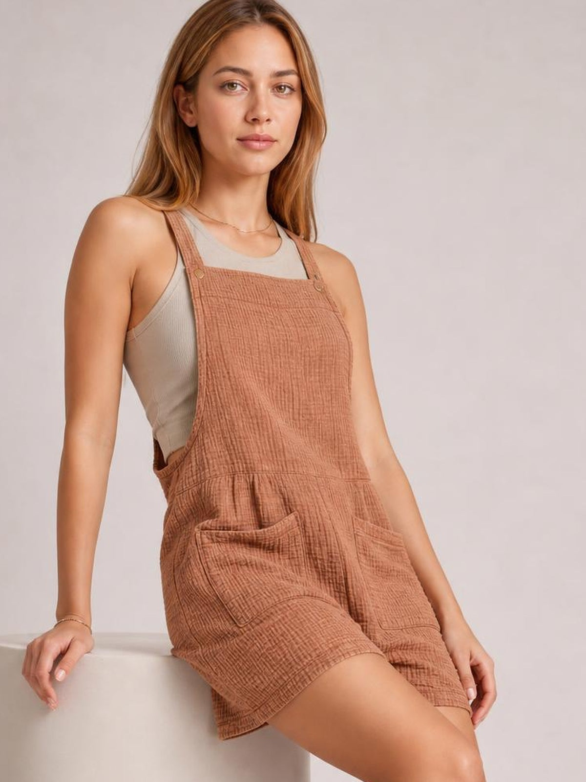 Model leans against a ledge wearing rust-brown sleeveless double gauze overalls with wide straps, a bib front, relaxed shorts, and large front pockets.