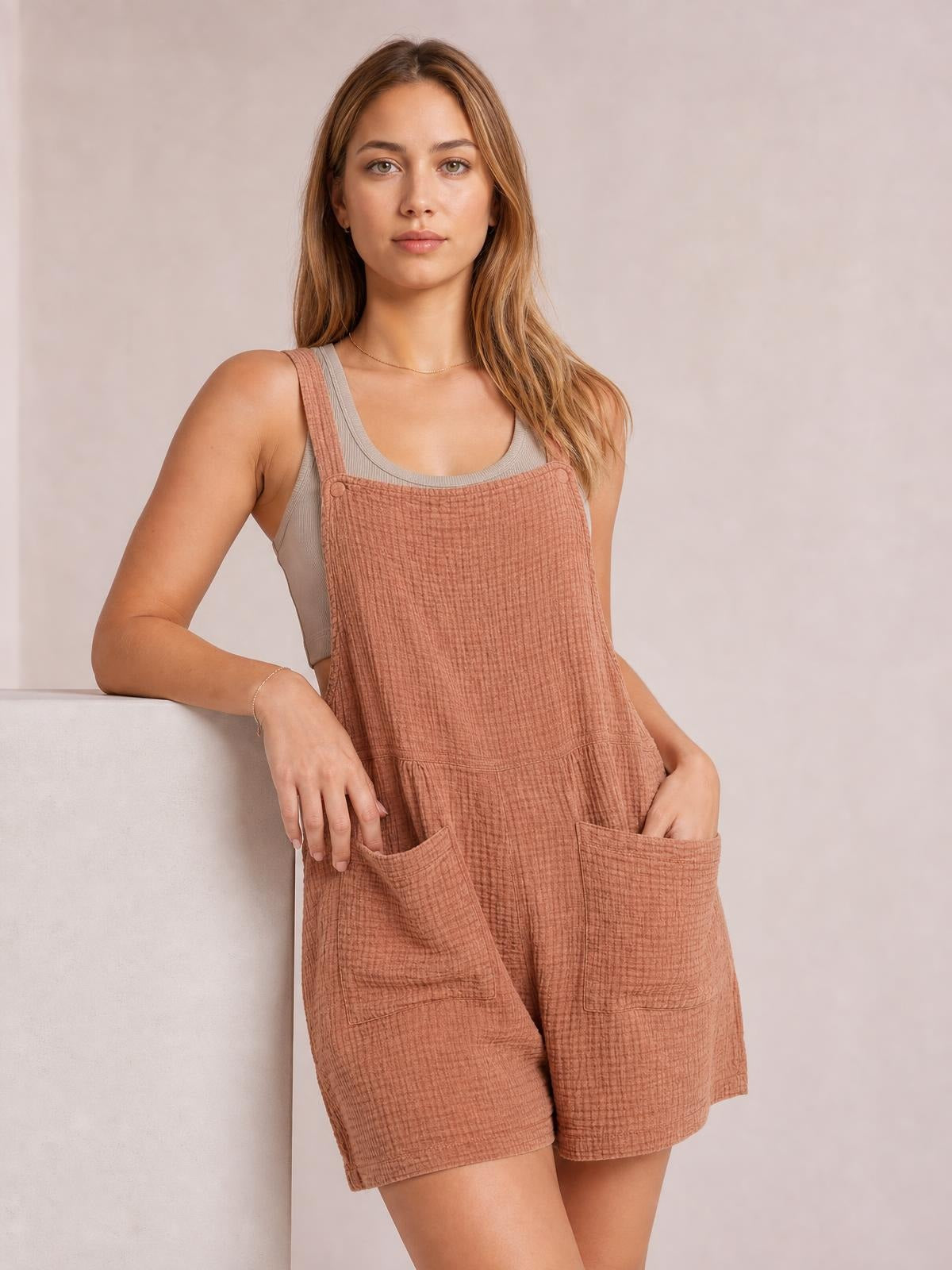 A model leans casually in a terracotta sleeveless double gauze overall romper with wide straps, a square neckline, relaxed shorts, and two front pockets.