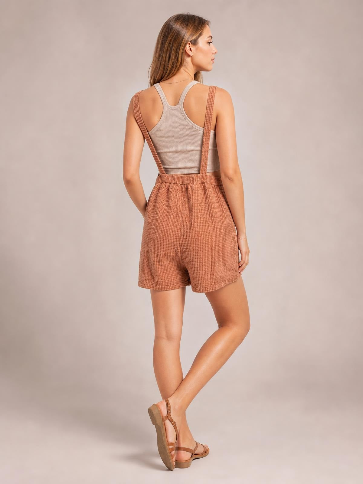 Model shown from behind in rust double gauze overalls with wide shoulder straps, relaxed shorts, textured fabric, and a hand tucked into a pocket.