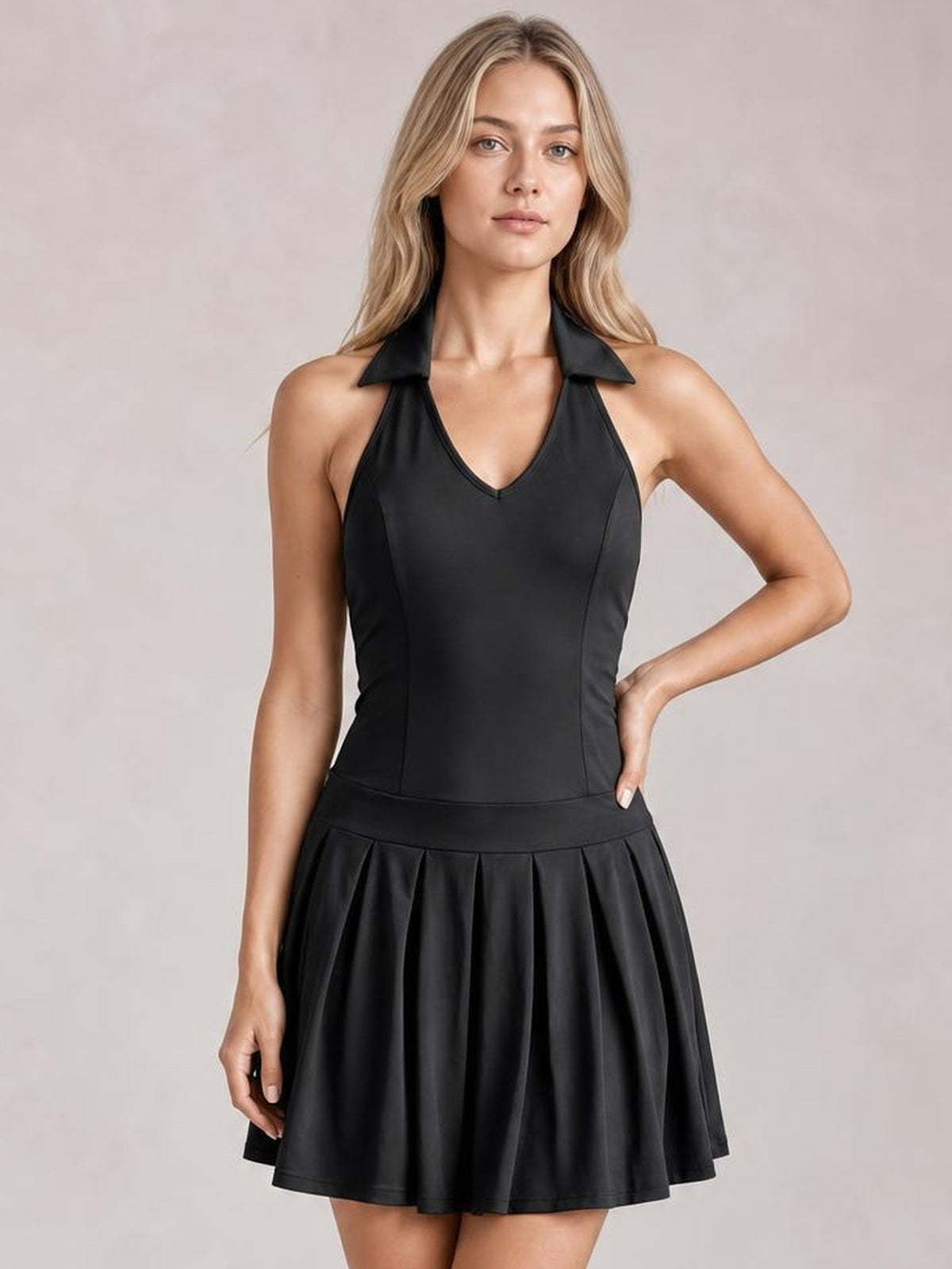 Model poses with one hand on her hip in a black sleeveless active dress featuring a fitted V-neck bodice, collared halter detail, and pleated flared skirt.