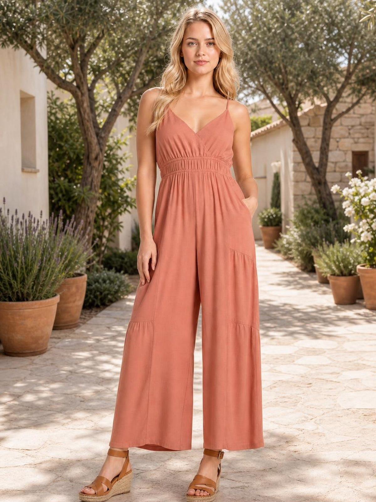 Model wearing a coral sleeveless ruched jumpsuit with adjustable spaghetti straps, elastic waist, tiered construction, wide legs, and a relaxed full-length silhouette.