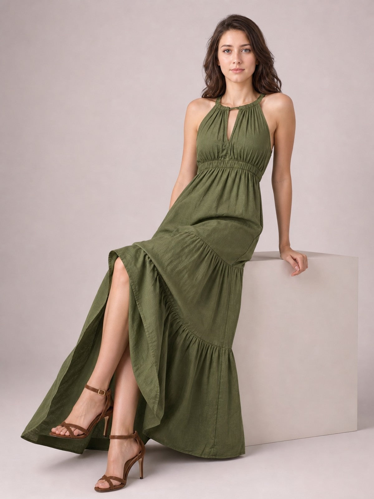 A seated model wears an olive green sleeveless tiered maxi dress with a halter neckline, keyhole front, smocked waist, and flowing gathered skirt.