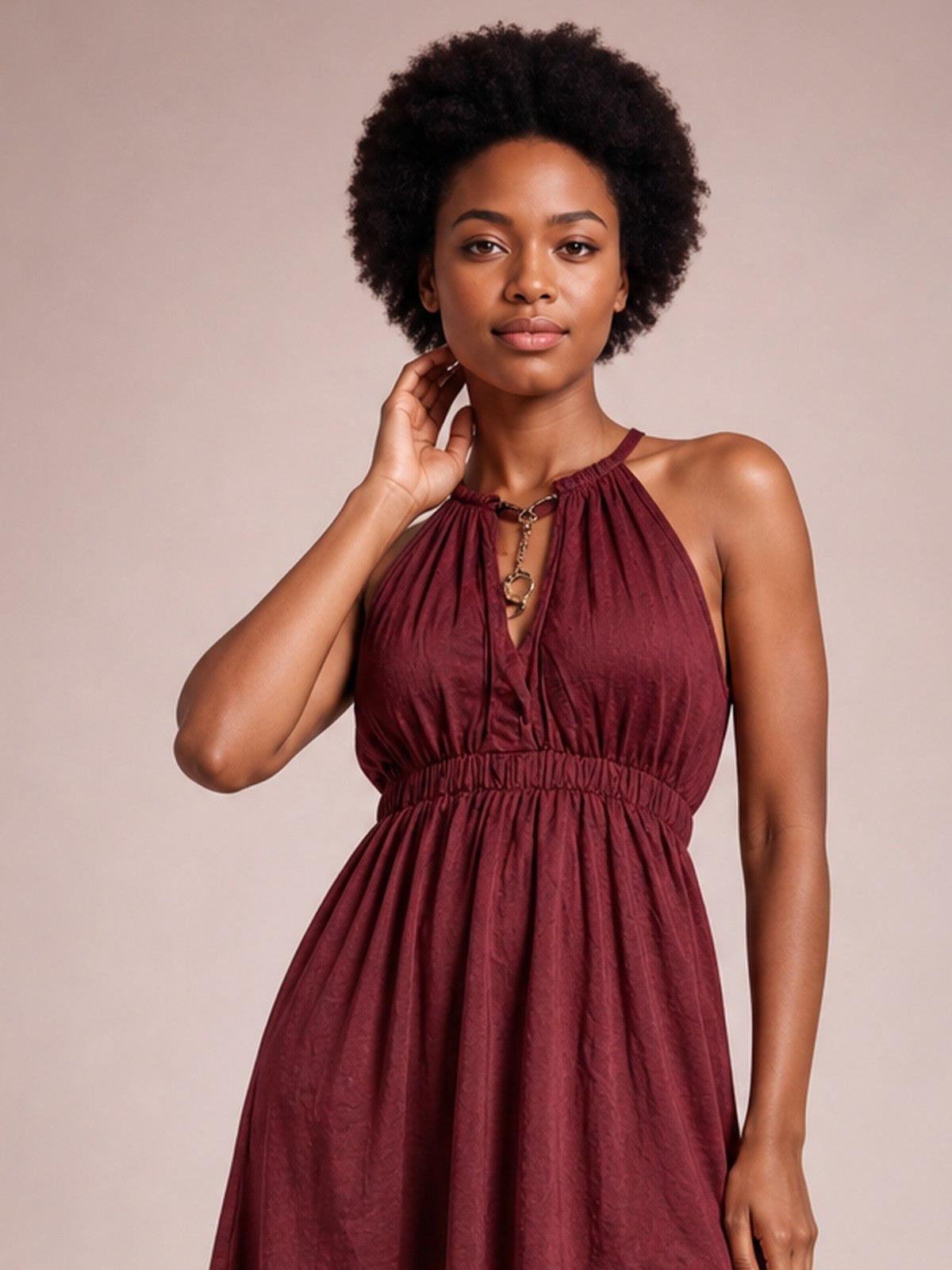 A model poses with one hand at her neck in a burgundy sleeveless maxi dress featuring a gathered halter neckline, keyhole detail, smocked waist, and flowing skirt.