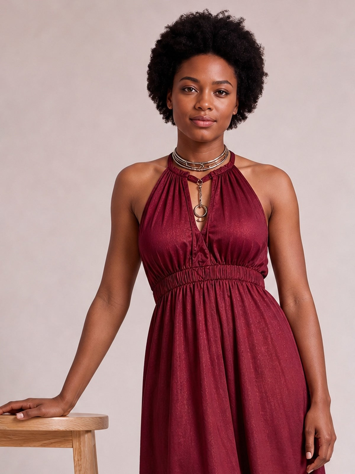 A model wears a burgundy sleeveless tiered maxi dress with a halter neckline, keyhole detail, gathered bodice, and smocked waist while resting one hand on a table.