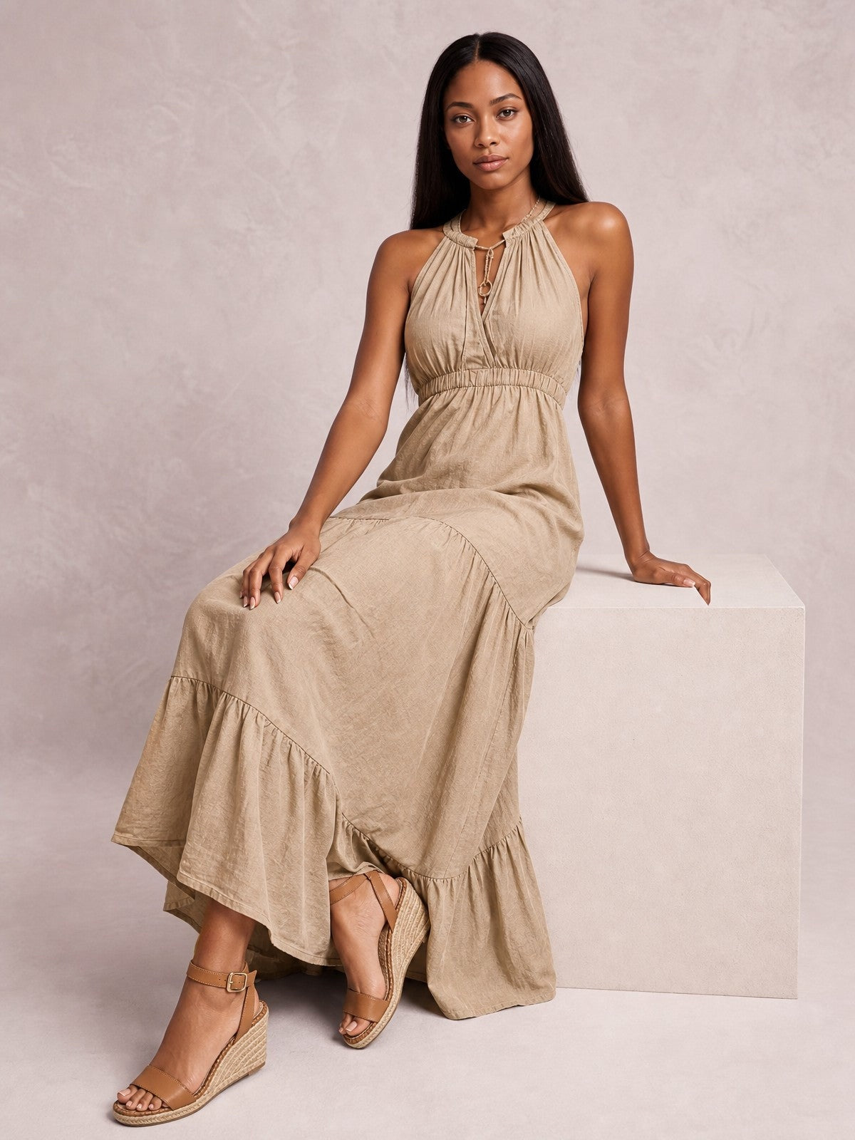 A seated model wears a beige sleeveless halter maxi dress with a gathered bodice, smocked waist, keyhole neckline, and flowing tiered skirt.