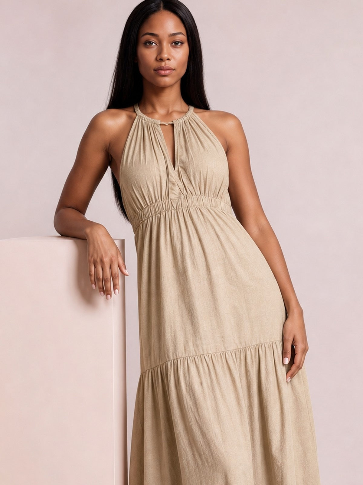 A model leans against a pedestal in a beige sleeveless halter maxi dress with a keyhole neckline, gathered bodice, smocked waist, and tiered skirt.