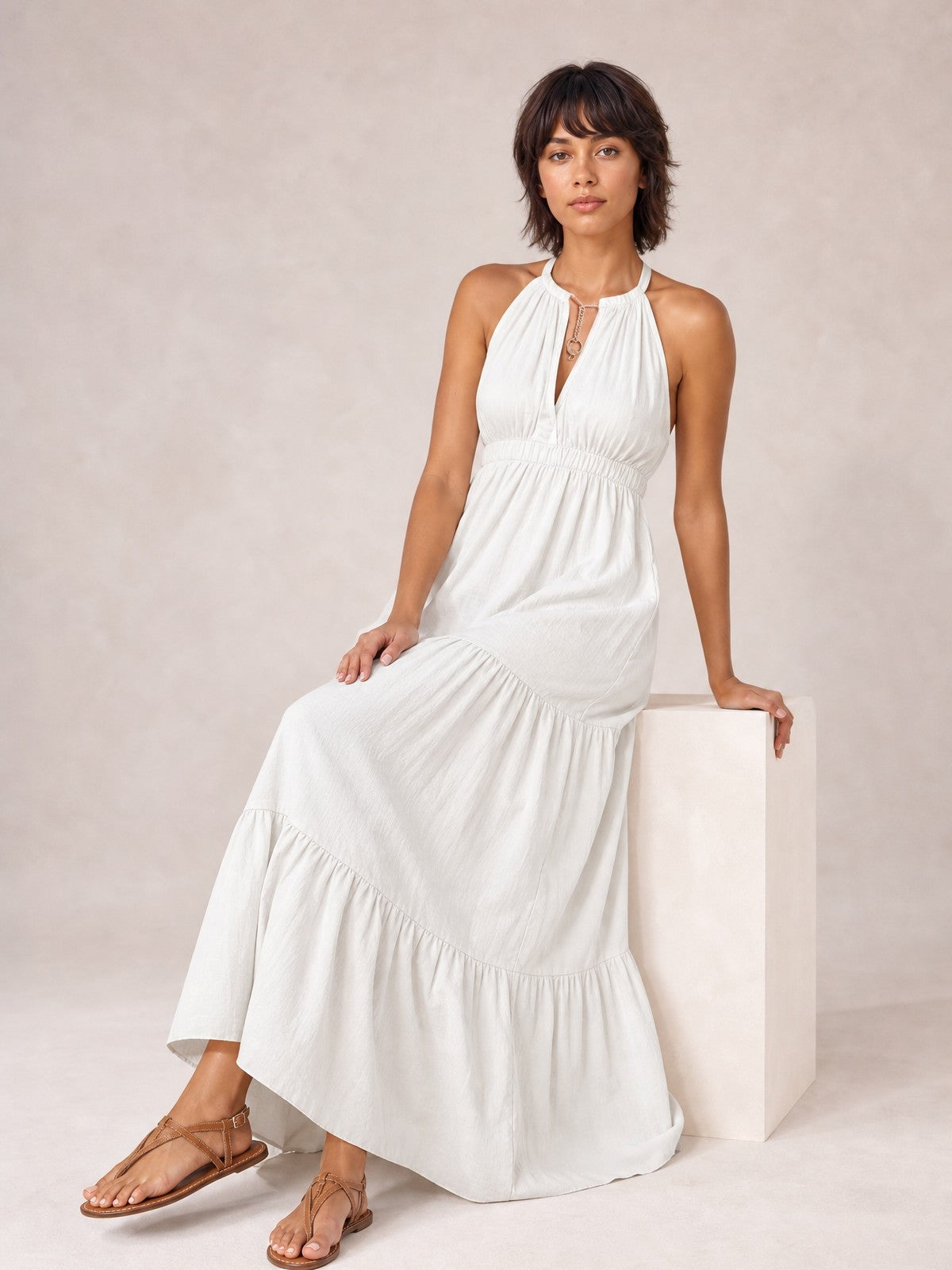 A model seated in a white sleeveless tiered maxi dress with a halter neckline, keyhole detail, gathered bodice, smocked waist, and flowing floor-length silhouette.