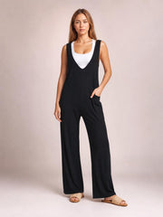 A model wears a black sleeveless wide leg jumpsuit with a deep V-neck bib, relaxed full-length silhouette, and front patch pockets, posing with one hand tucked inside.