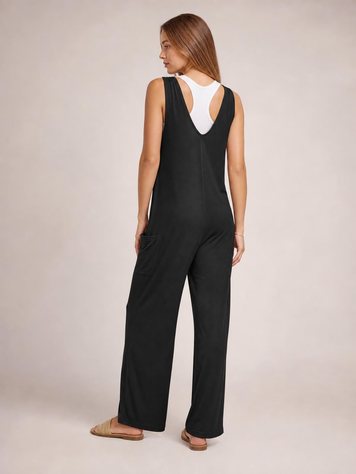Model shown from behind in a black sleeveless wide-leg jumpsuit with a deep V-back, center seam, and side pocket, standing with one hand near the pocket.