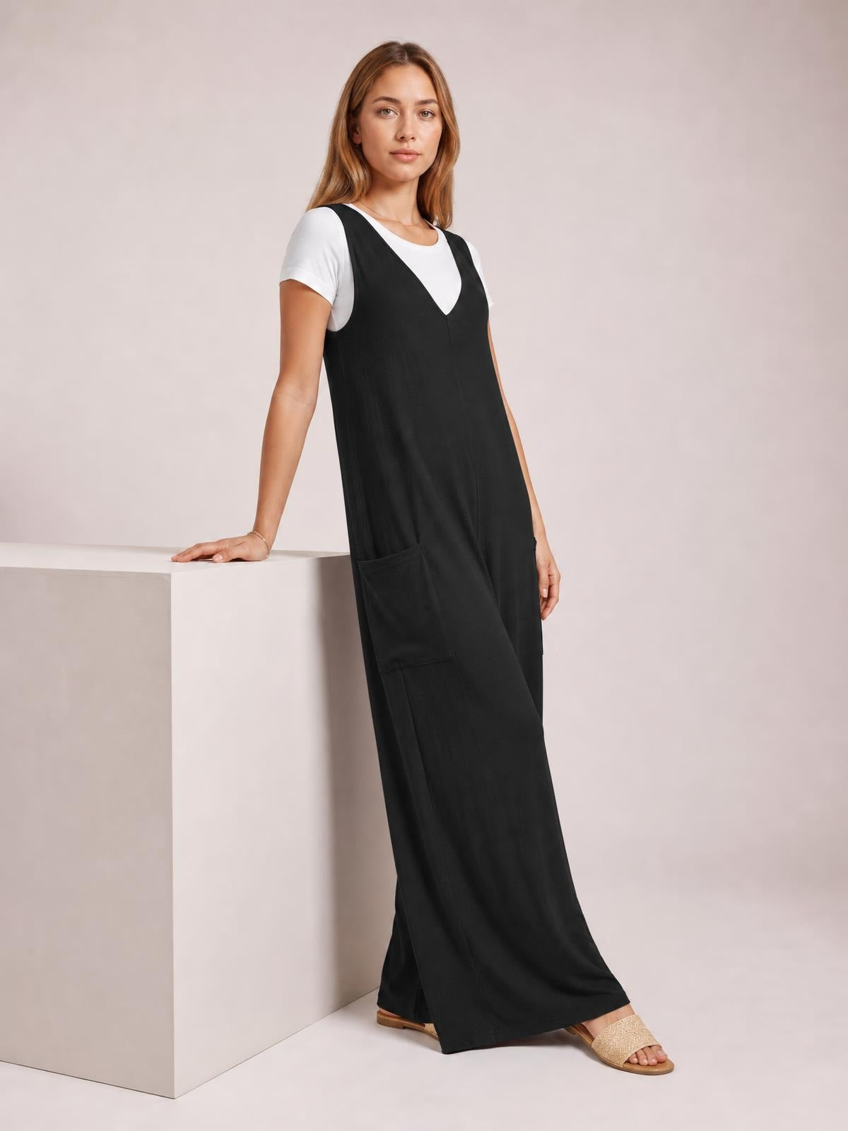 Model wears a black sleeveless wide-leg jumpsuit over a white tee, featuring a V-neckline, relaxed silhouette, and large front pockets.