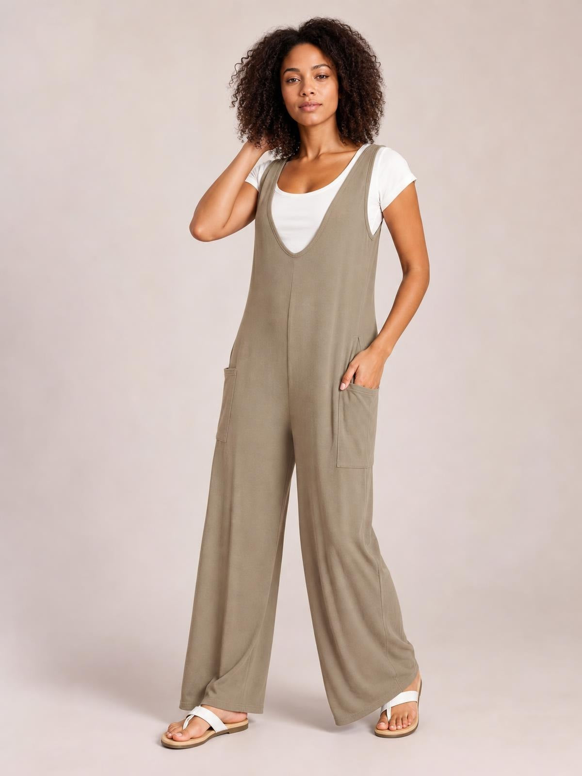 A model poses in a taupe sleeveless wide leg jumpsuit layered over a white tee, featuring a deep V neckline and large patch pockets.