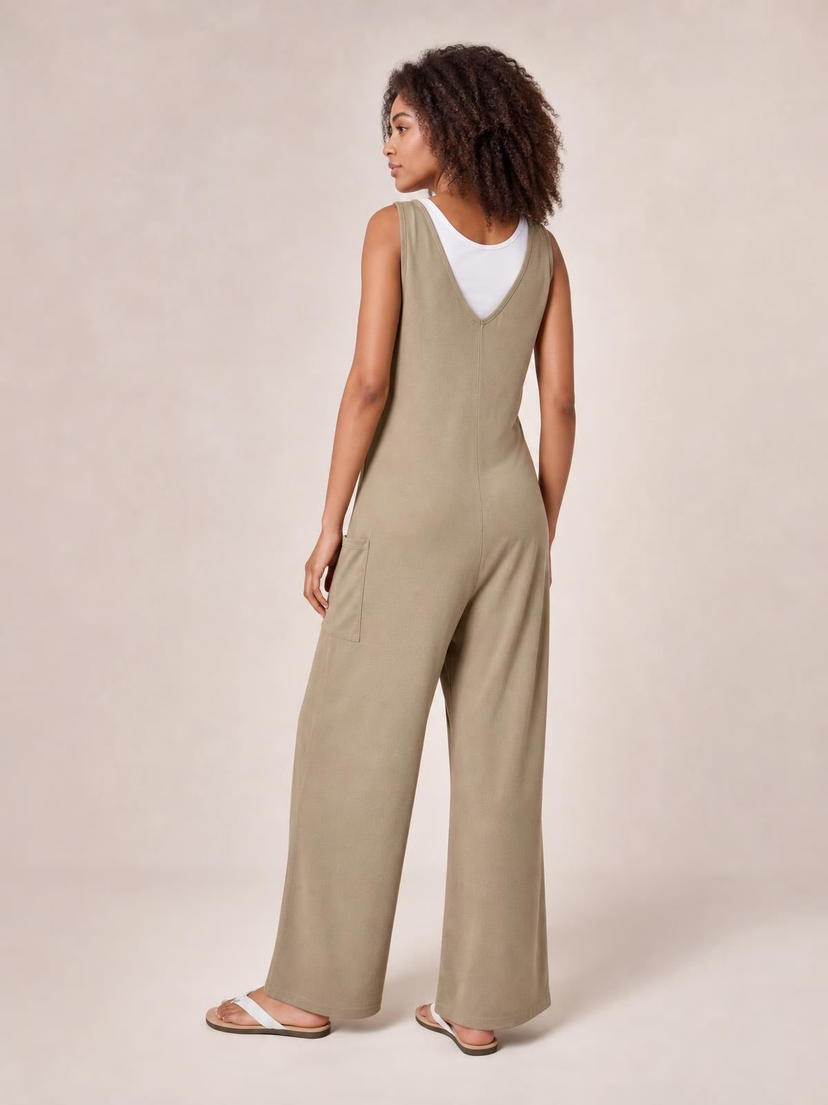 A model looks over her shoulder in a taupe sleeveless wide-leg jumpsuit with a deep V-back, relaxed silhouette, and visible patch pocket.