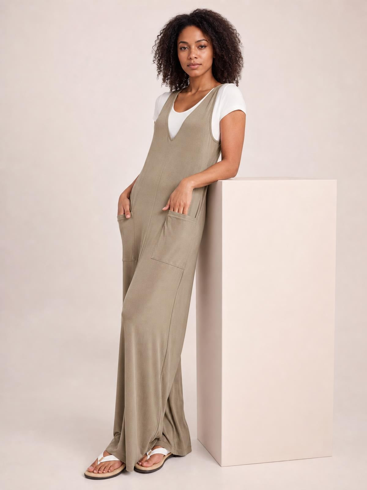 A model leans beside a pedestal wearing a taupe sleeveless wide-leg jumpsuit with a relaxed silhouette, V-neckline, and two front pockets.