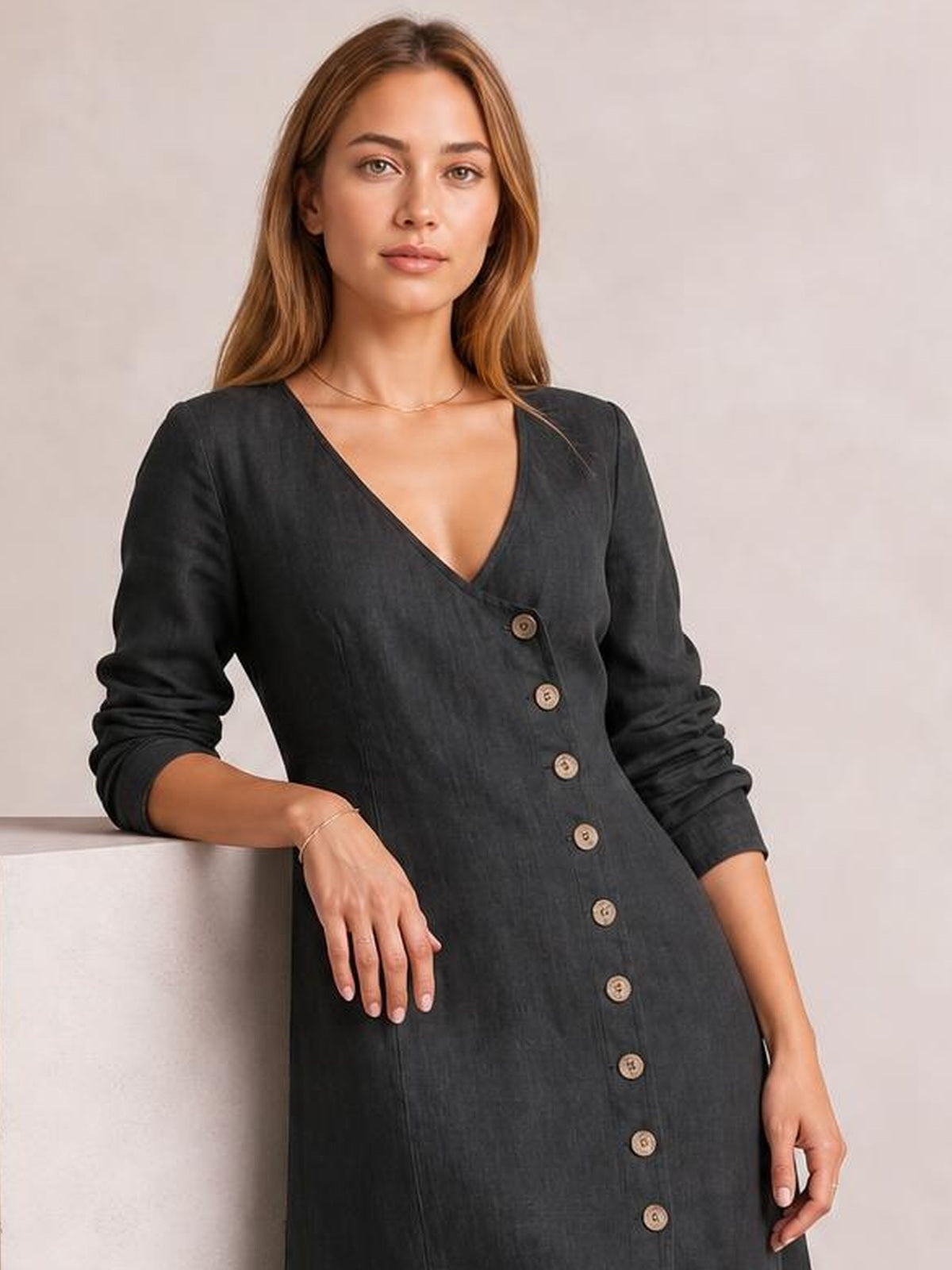 Model wears a dark gray button-down midi dress with a V-neckline, long sleeves, and front button placket while leaning with one hand on a ledge.