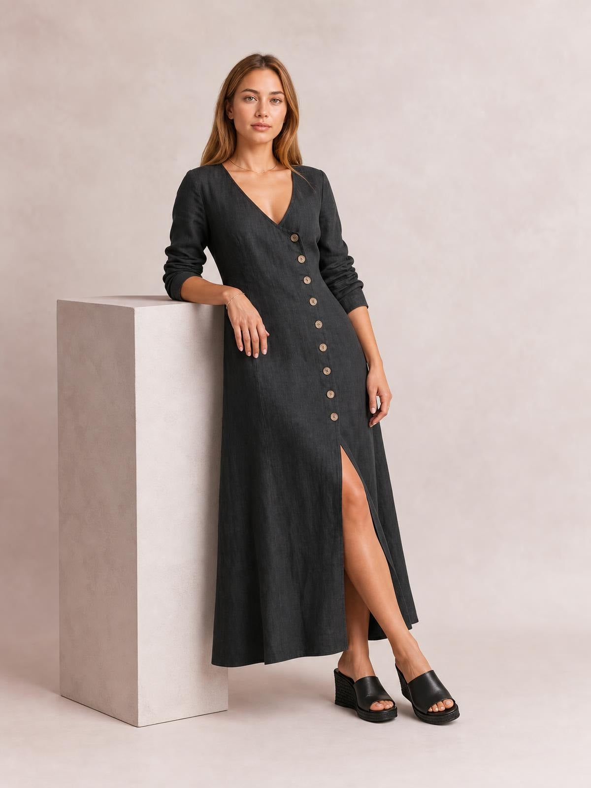 Model wearing a black button-down midi dress with a V-neckline, long sleeves, relaxed straight silhouette, and high front slit while leaning against a pedestal.