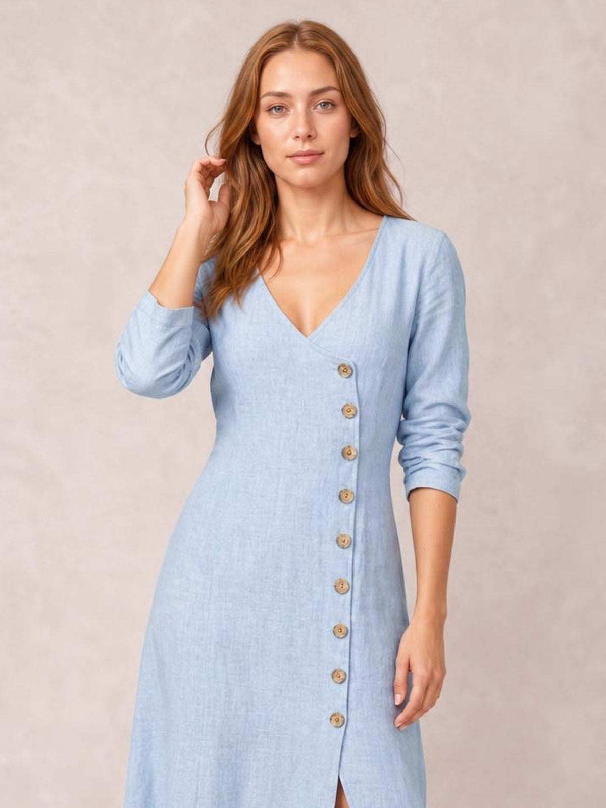 Model poses in a light blue button-down midi dress with a V-neck, long sleeves, fitted waist, front button construction, and lower slit.