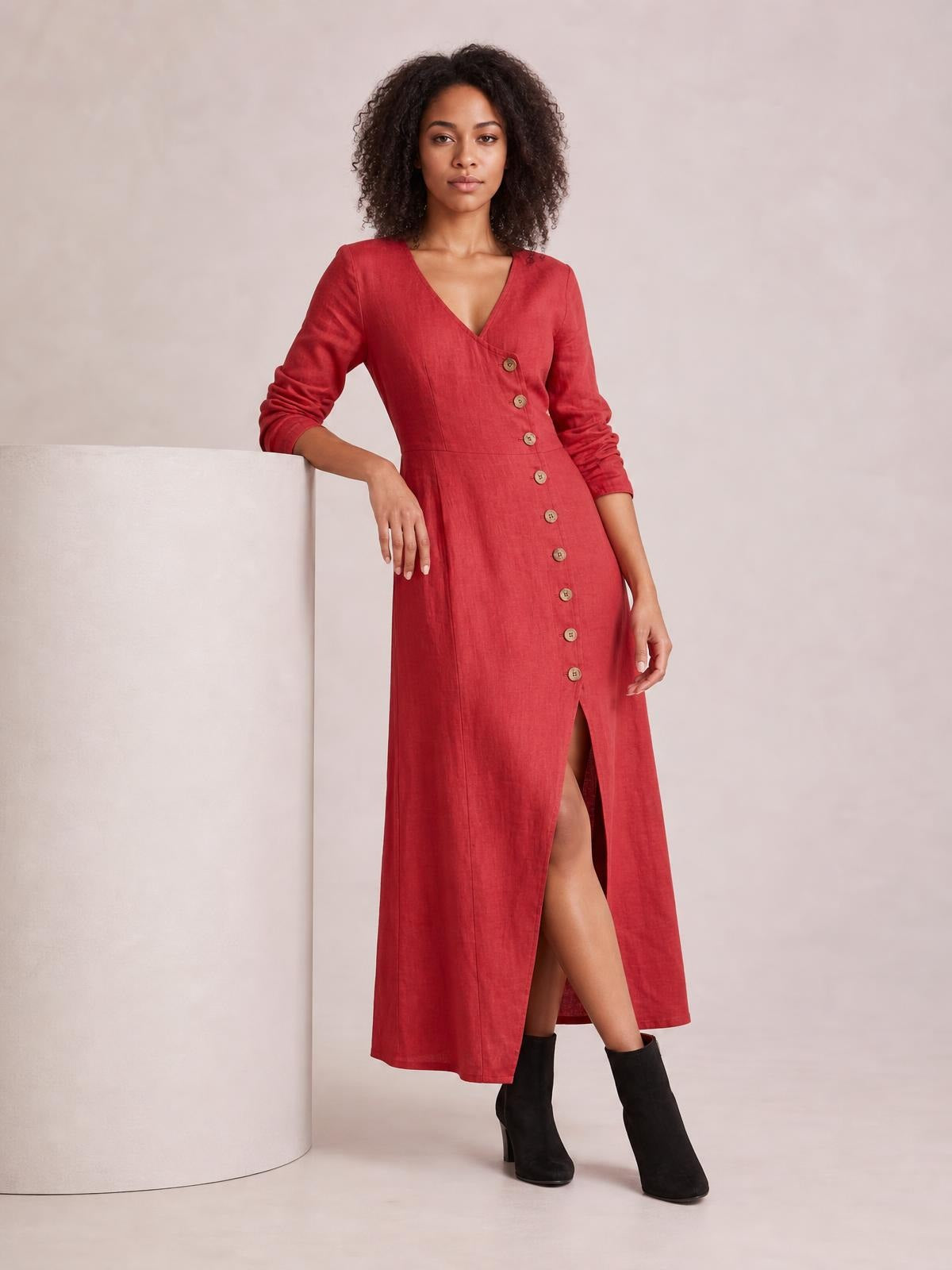 A model leans against a pedestal wearing a red button-down midi dress with long sleeves, a V-neck, shaped waist, straight silhouette, and front slit.