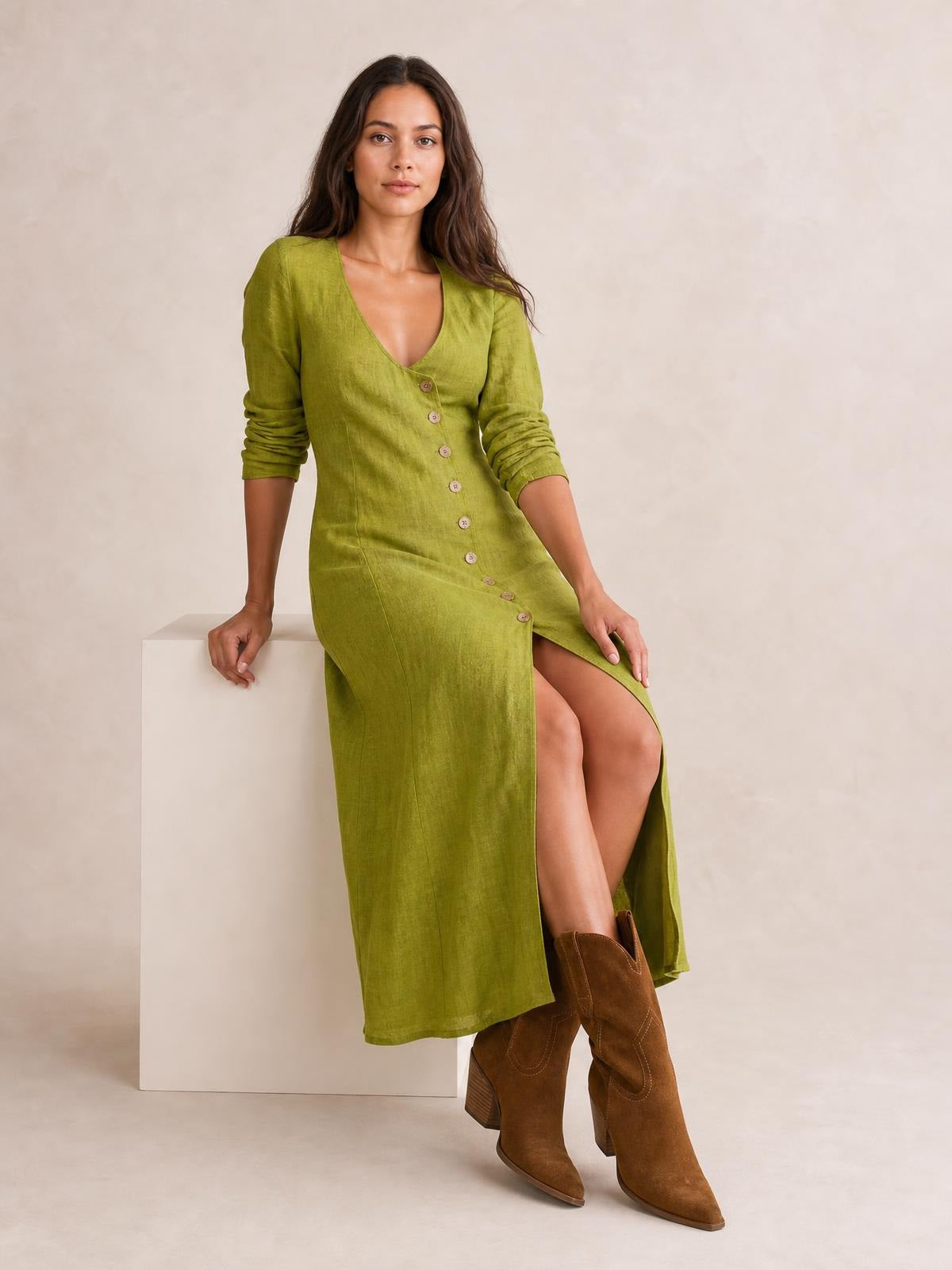 Model wears an olive green button-down midi dress with a V-neck, long ruched sleeves, slim silhouette, and high side slit, seated with one leg crossed.