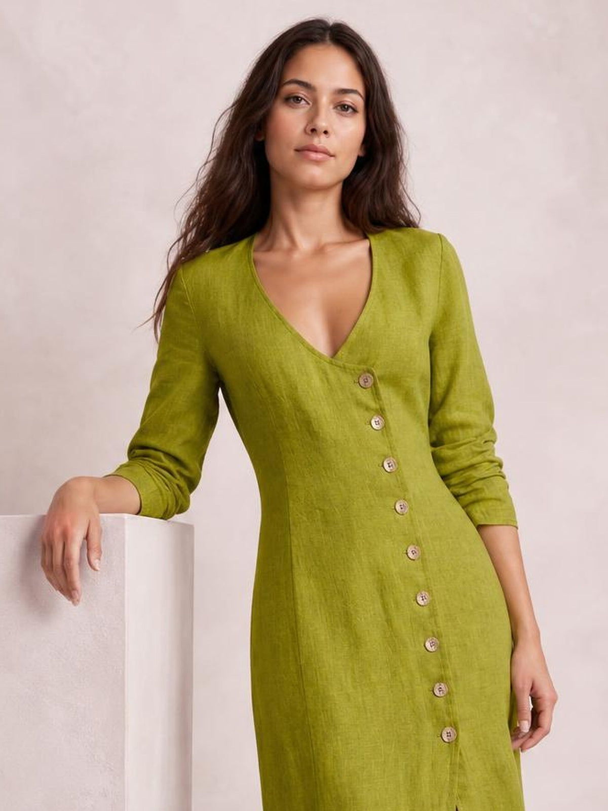 Model wears an olive-green button-down midi dress with a V-neck, long sleeves, straight silhouette, and visible front button construction while leaning on a pedestal.