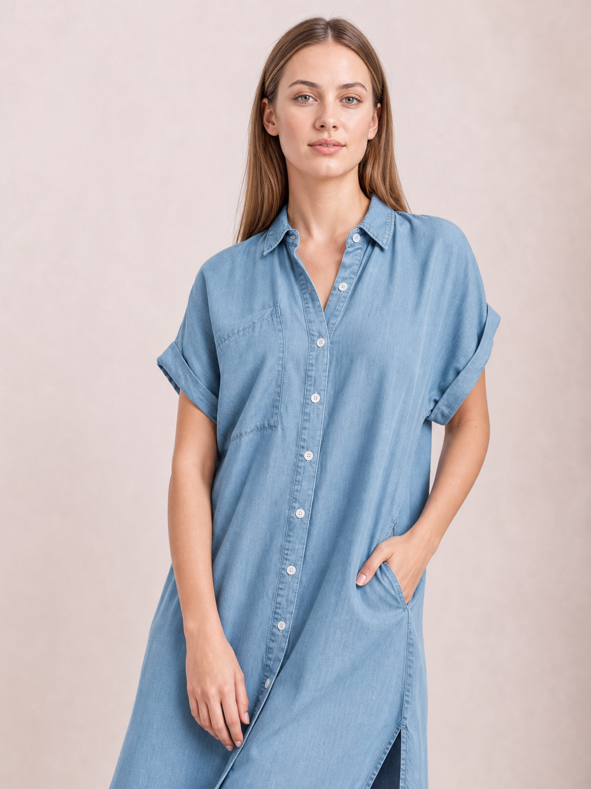 Model poses with one hand in the pocket of a light denim-blue button-up dress featuring a classic collar, rolled short sleeves, straight silhouette, and side slit.