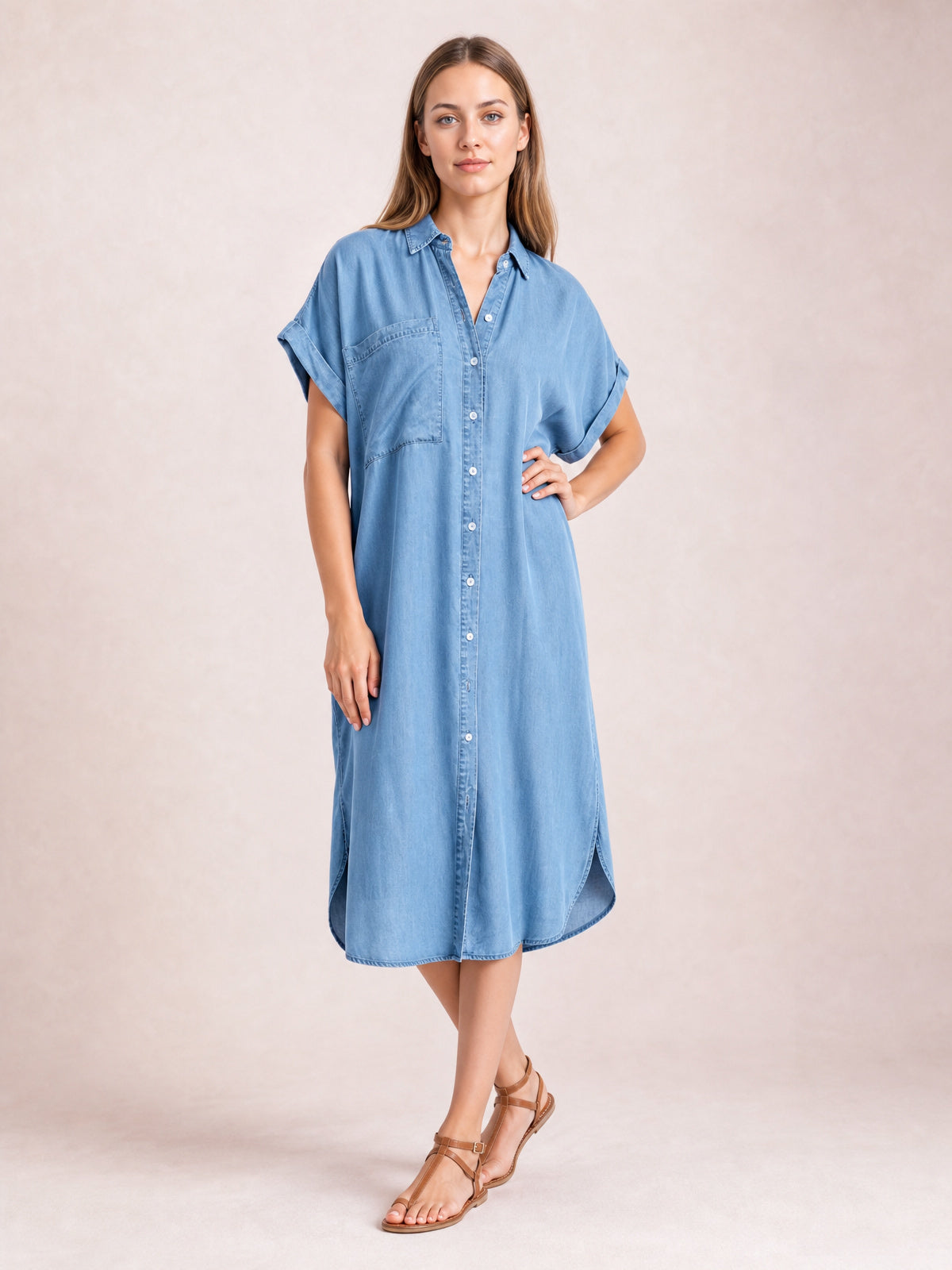 Model poses in a medium denim-blue button-up midi dress with a collar, short rolled sleeves, straight silhouette, front pocket, and side slits.