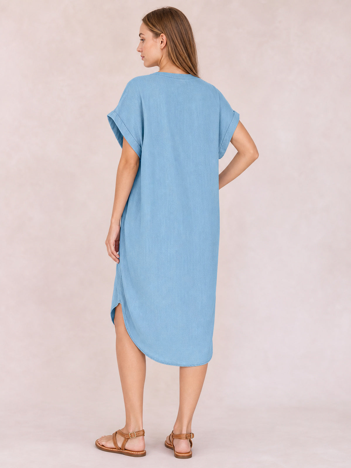 Model shown from the back wearing a medium blue imitation denim dress with short rolled sleeves, a straight midi silhouette, curved hem, and side slit.