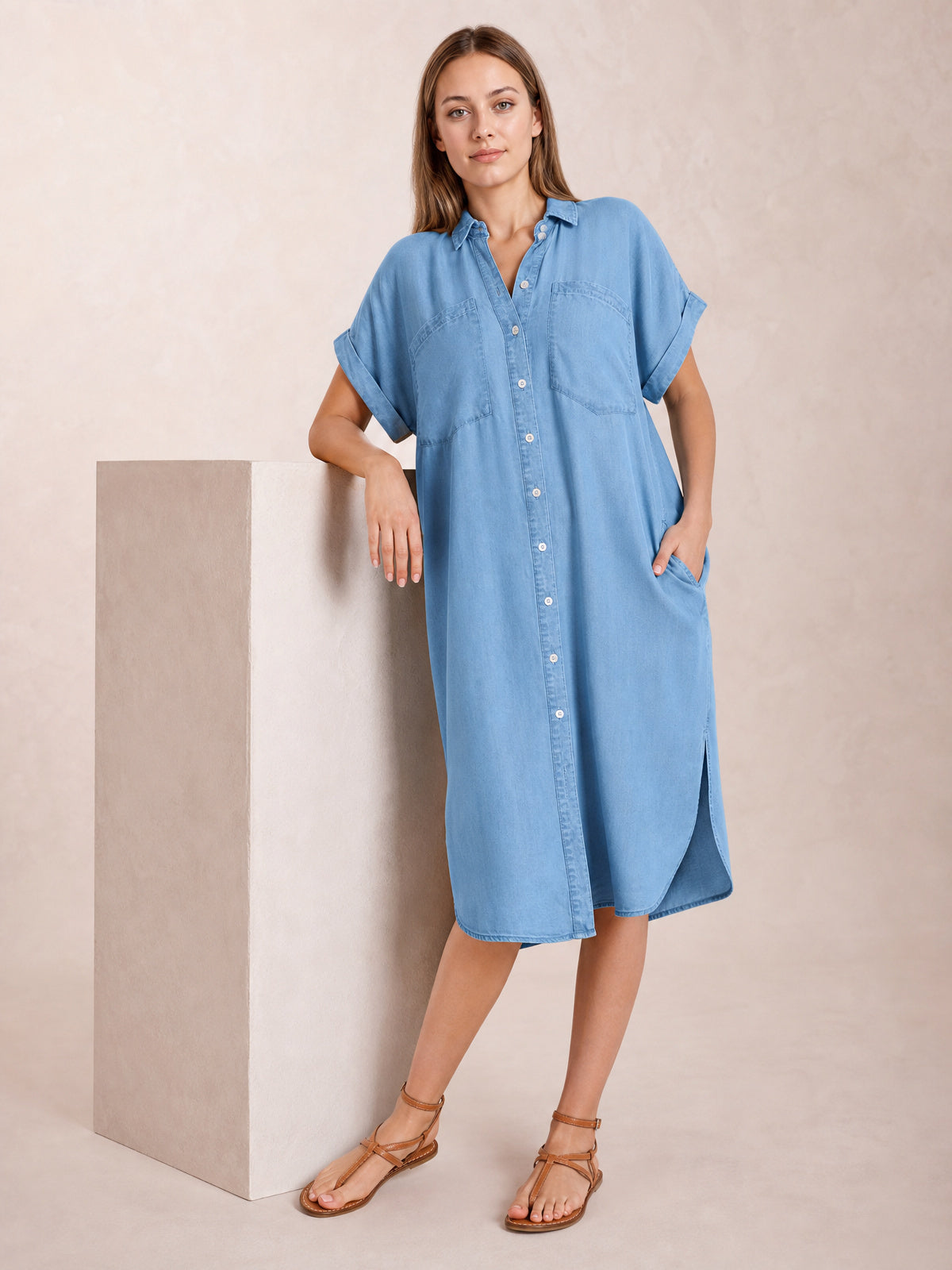 Model poses in a medium denim-blue button-up midi dress with a collar, rolled short sleeves, chest pockets, relaxed straight silhouette, and side slits.