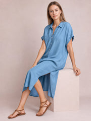 Model seated in a light denim-blue imitation denim dress with a button-front, classic collar, short rolled-cuff sleeves, straight midi silhouette, and side slit.
