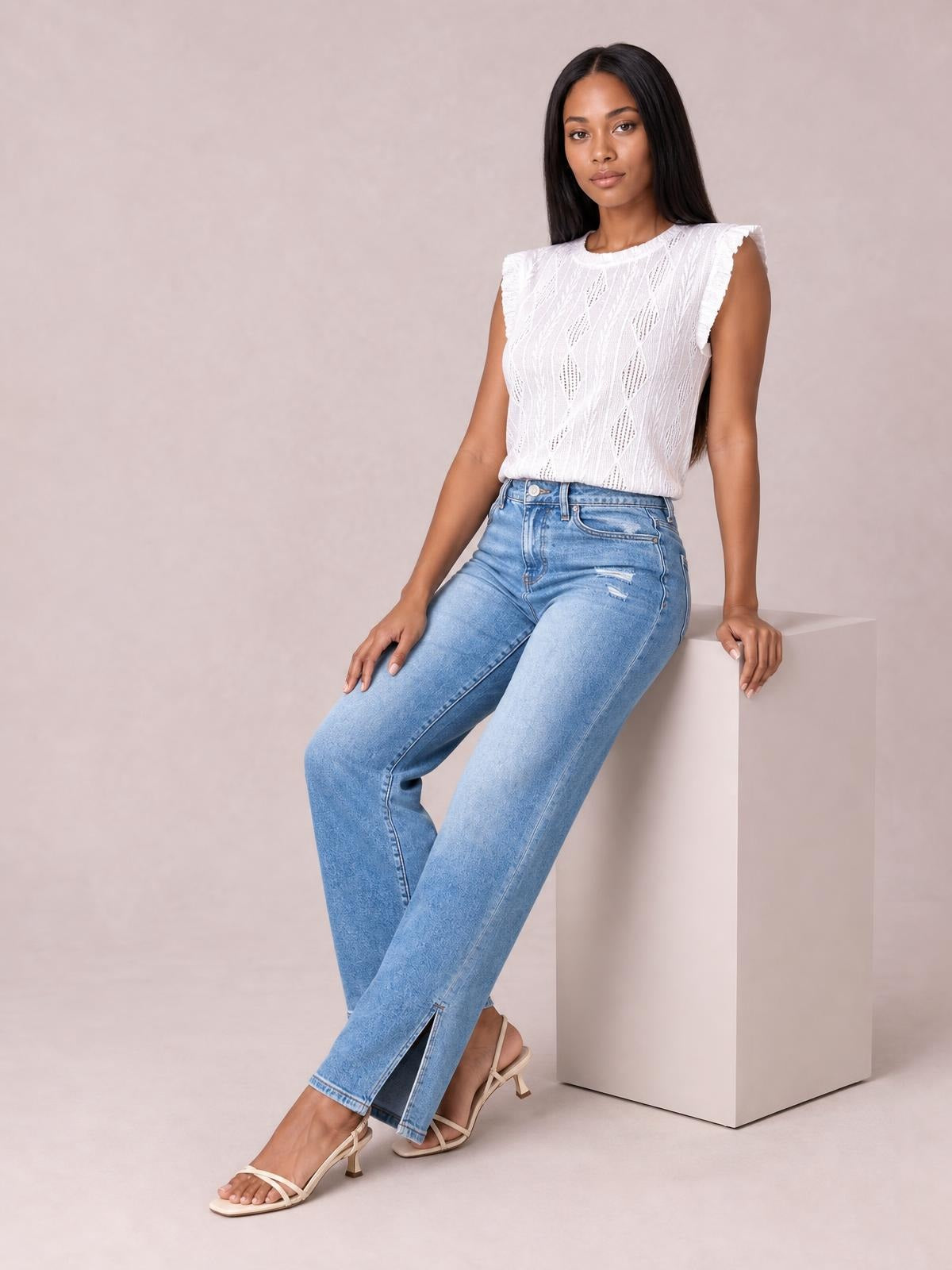 A seated model wears light-wash blue jeans with a buttoned waist, front pockets, a slim straight-leg silhouette, and visible ankle slits.