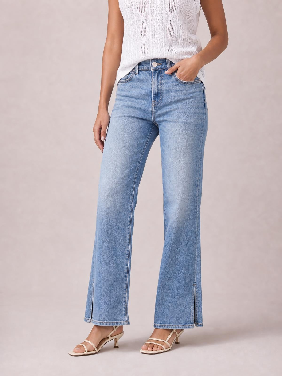 A model wears light-wash jeans with a button fly, front pockets, straight wide legs, and ankle-length side slits, posing with one hand in a pocket.