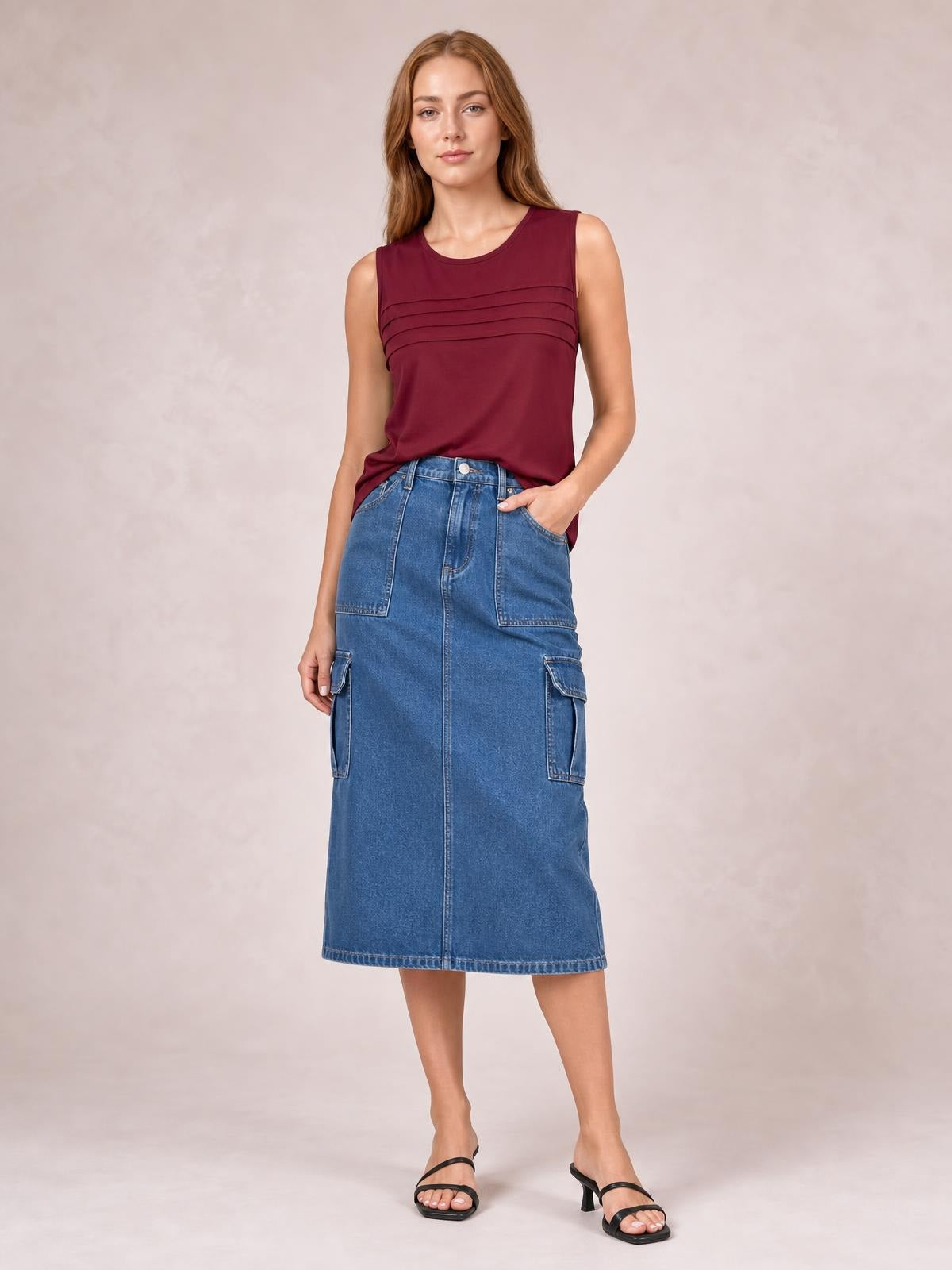 A model poses in a blue denim midi skirt with a straight silhouette, front seam, waistband, belt loops, and utility pockets, resting one hand in a pocket.