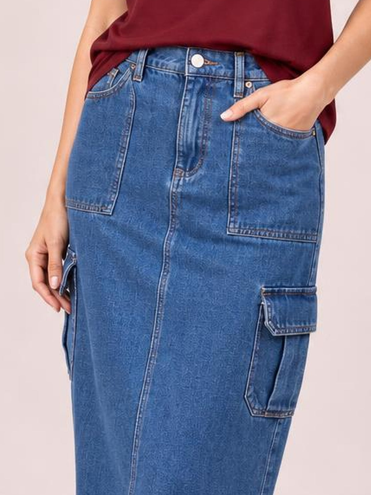 A model wears a blue denim midi skirt with a straight silhouette, button fly, belt loops, stitched front pockets, and roomy cargo-style patch pockets.
