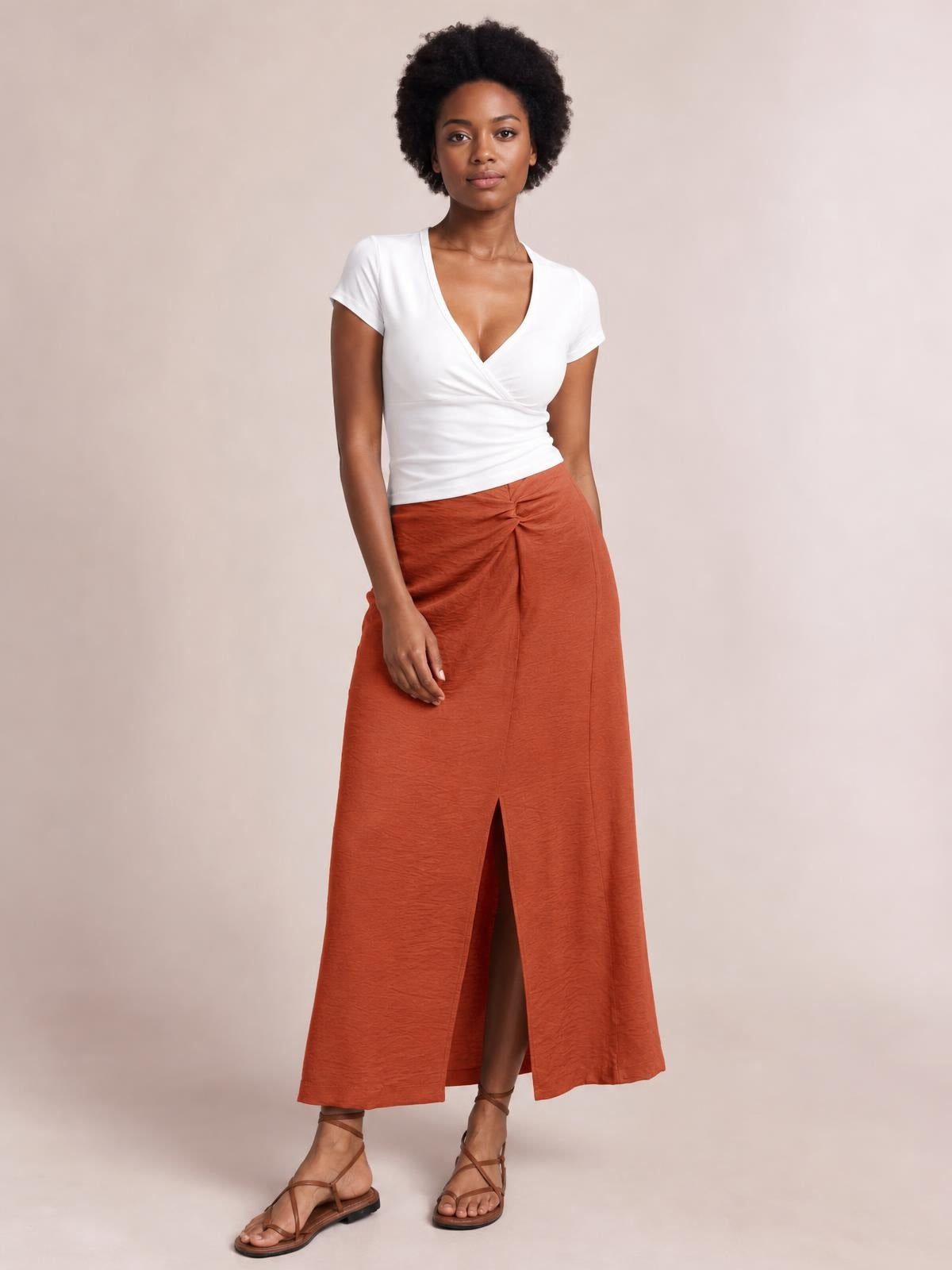 Model wearing a rust-orange Slit Midi Skirt with a flowy A-line silhouette, front slit, and gathered waist detail, styled with a white wrap top.