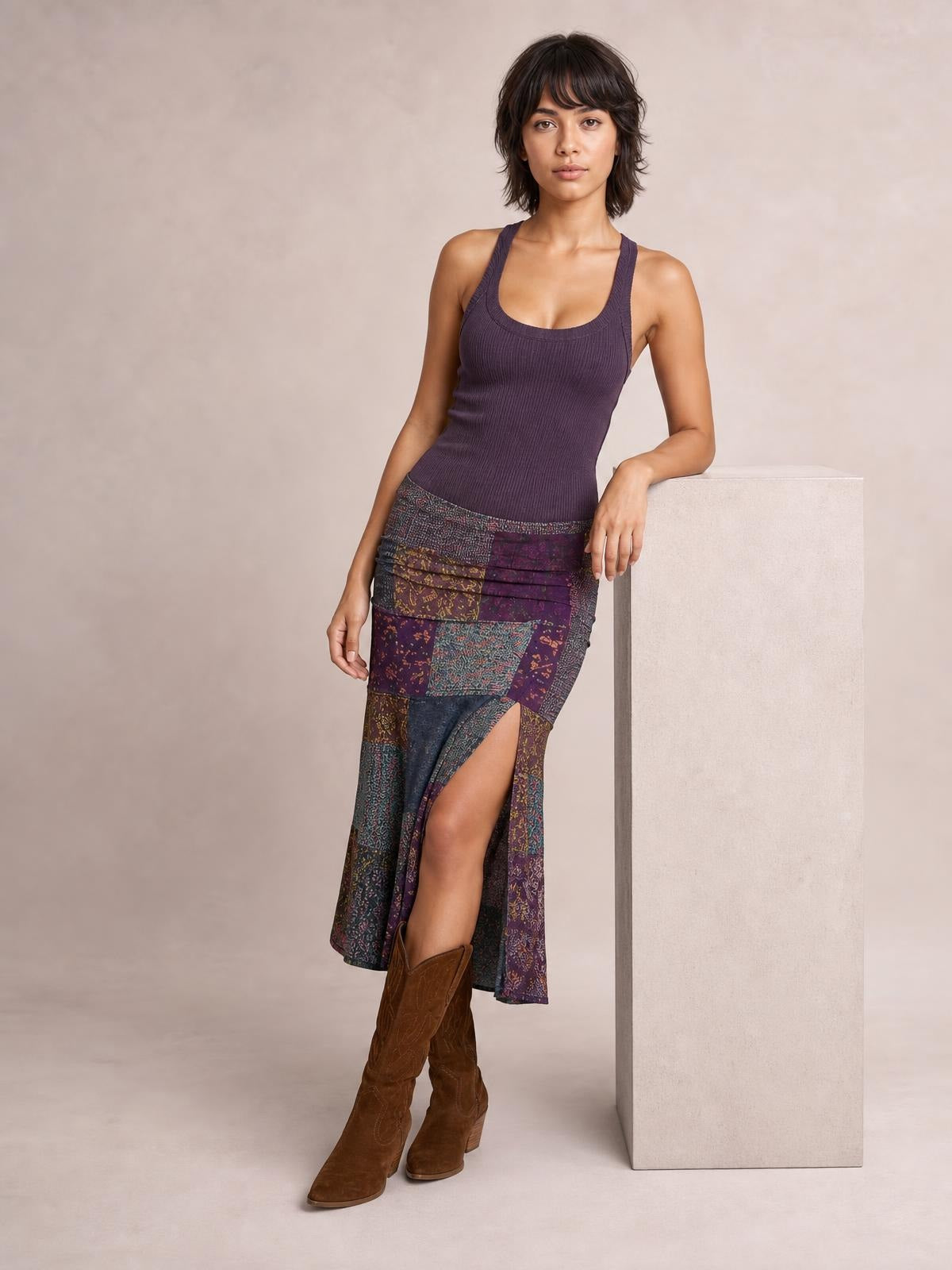 A model leans against a block in a fitted sleeveless midi dress with a purple ribbed bodice, colorful printed patchwork skirt, and thigh-high split.