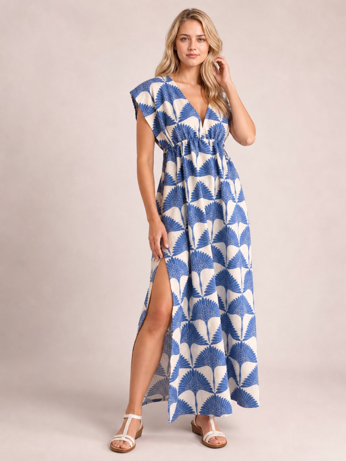 Model poses in a blue-and-white printed V-neck cap-sleeve maxi dress with a ruched waist, flowing silhouette, and thigh-high side slit.