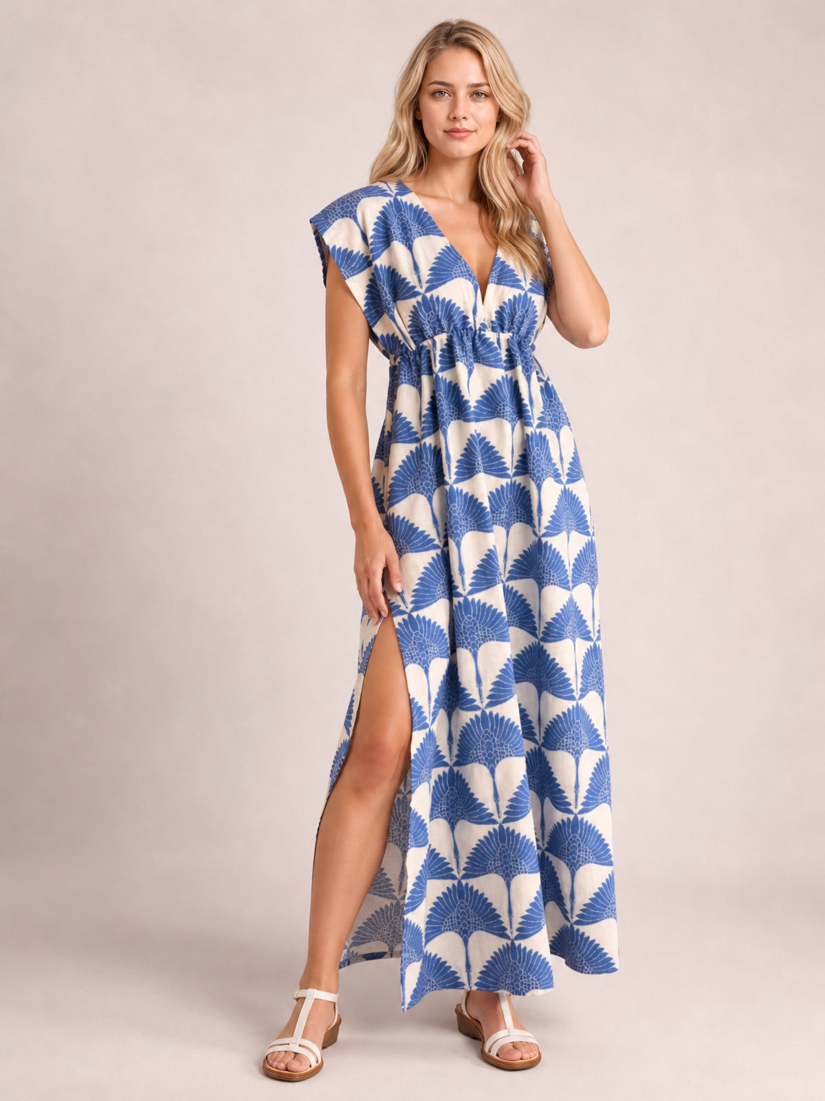 Model poses in a blue-and-white printed V-neck cap-sleeve maxi dress with a ruched waist, flowing silhouette, and thigh-high side slit.