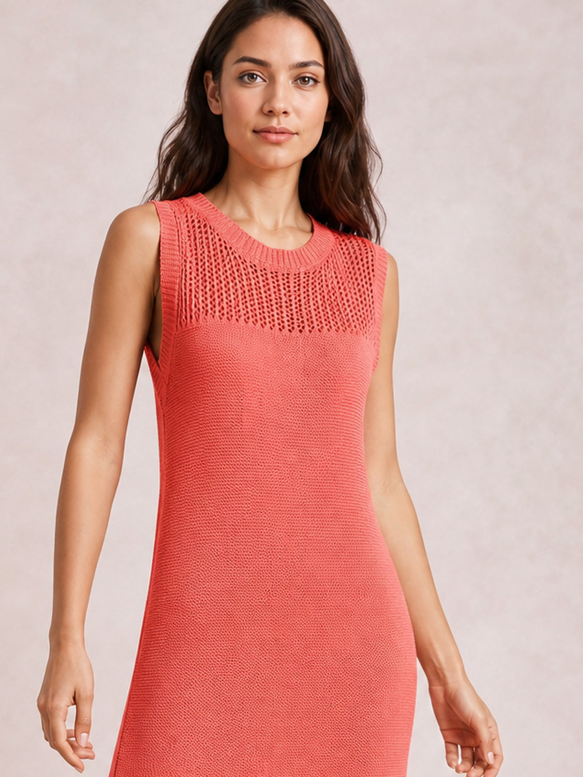 Model wearing a coral sleeveless sweater dress with a round neck, openwork knit panel, fitted elongated silhouette, and side slit detail.